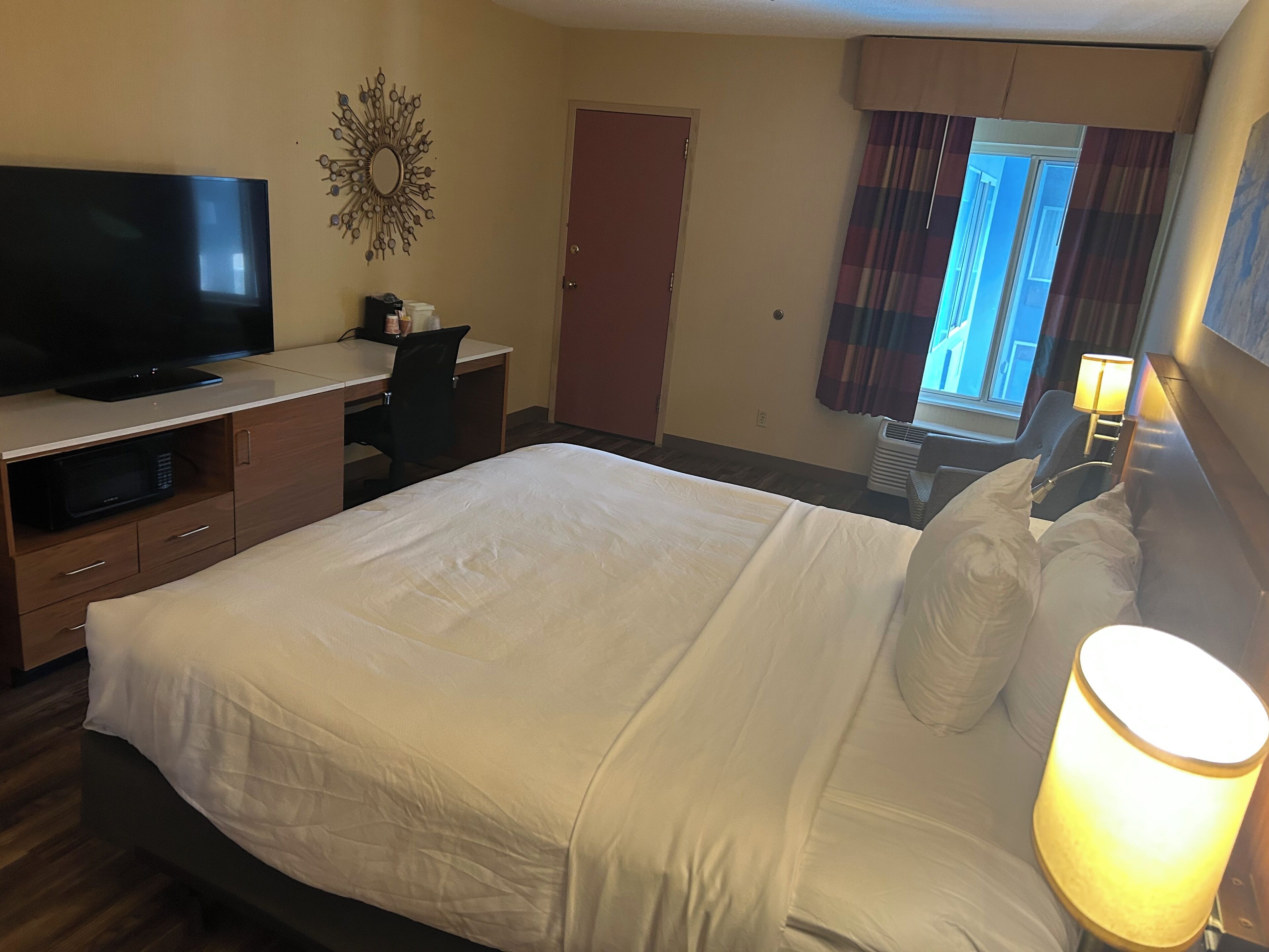 Ein Hotelzimmer mit Bett, Schreibtisch mit Fernseher, Spiegel, Fenster mit Vorhängen und Lampe.