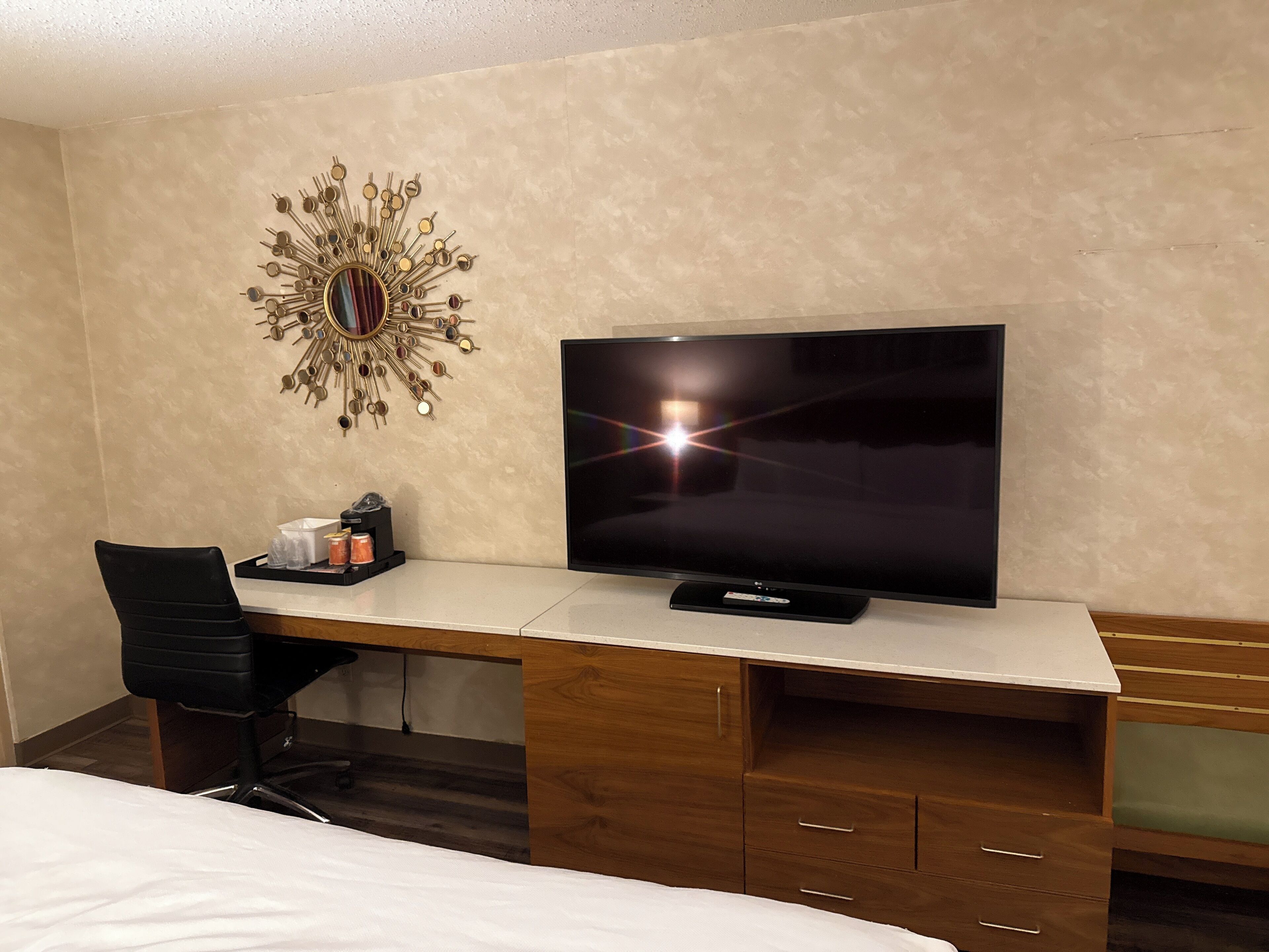 Ett hotellrum med ett skrivbord i trä, en platt-TV, en dekorativ väggdekoration, en stol och en säng.