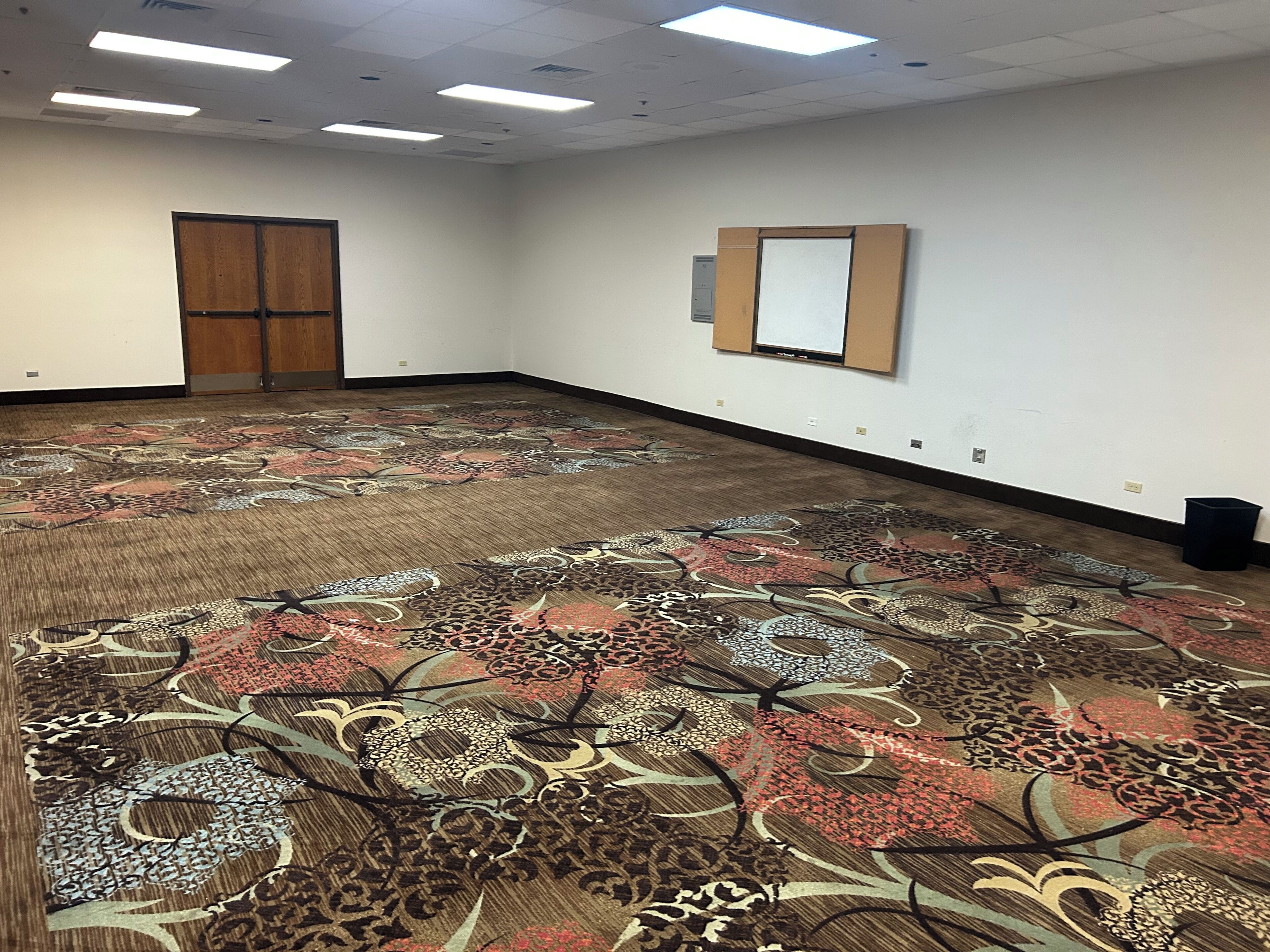 Un salón de conferencias amplio y vacío, con alfombra estampada, una puerta de madera, una pizarra y un bote de basura negro.