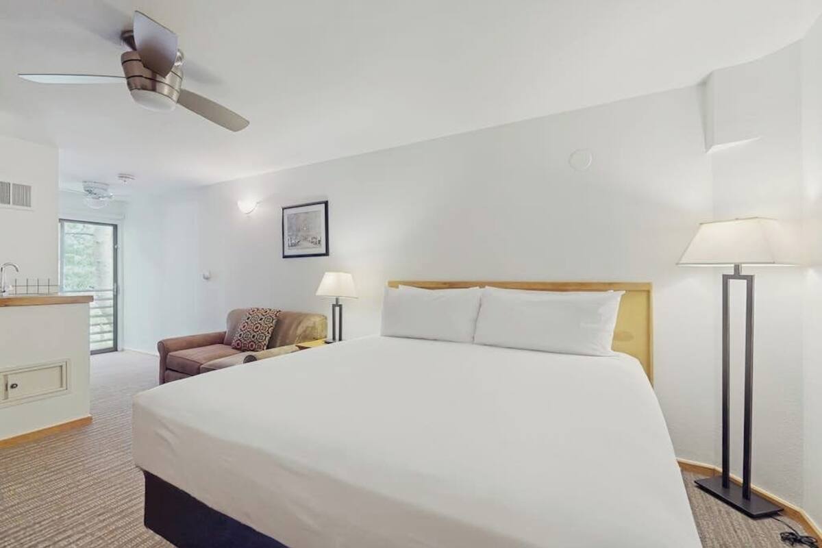 A hotel room with a large bed, a desk, a chair, a standing lamp, and a ceiling fan.