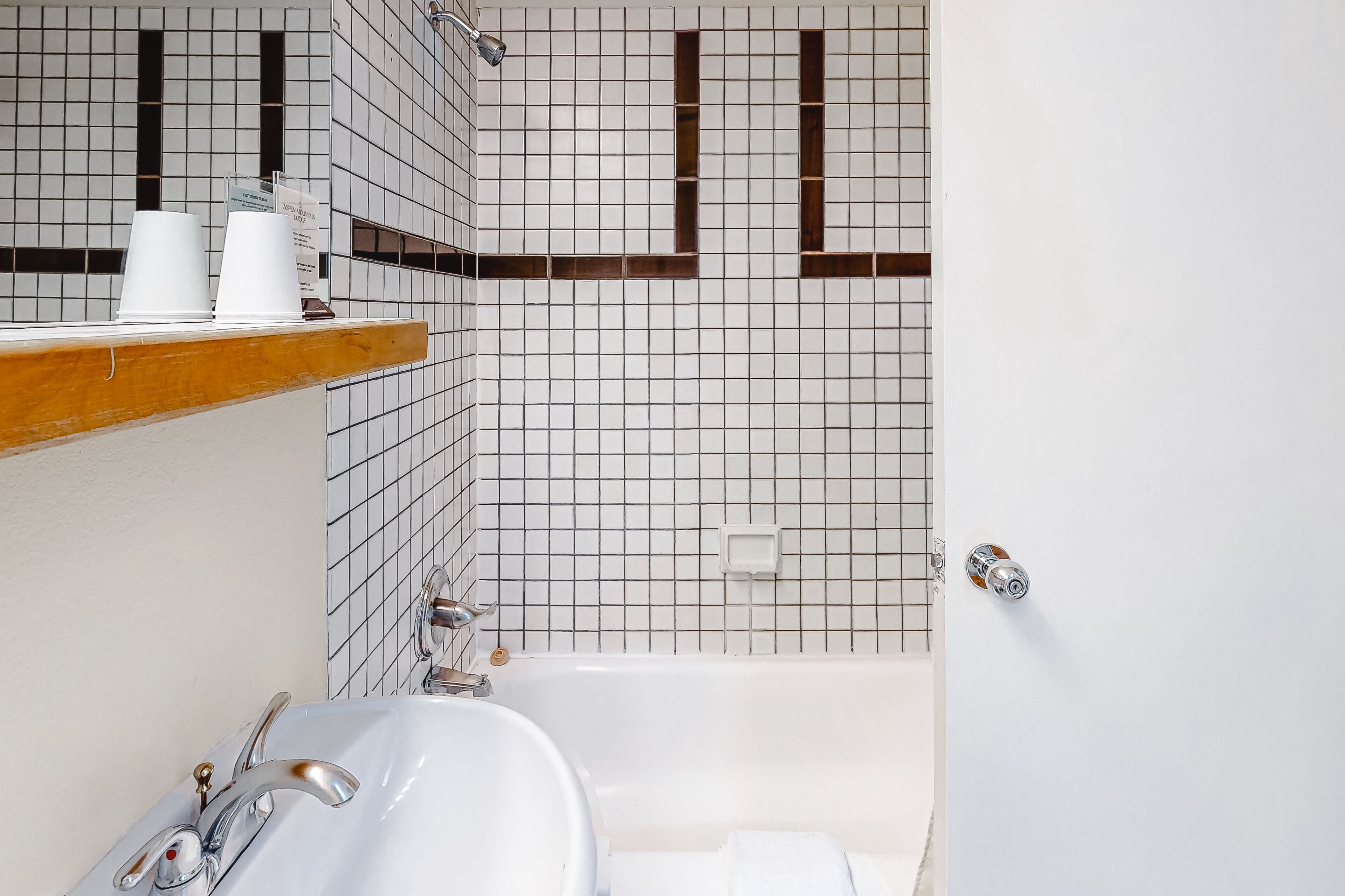Un baño con lavamanos blanco, espejo y bañera. Hay dos tazas blancas sobre una repisa de madera sobre el lavamanos. Las paredes están cubiertas de azulejos con un diseño de cuadrícula y presentan algunas líneas verticales marrones.