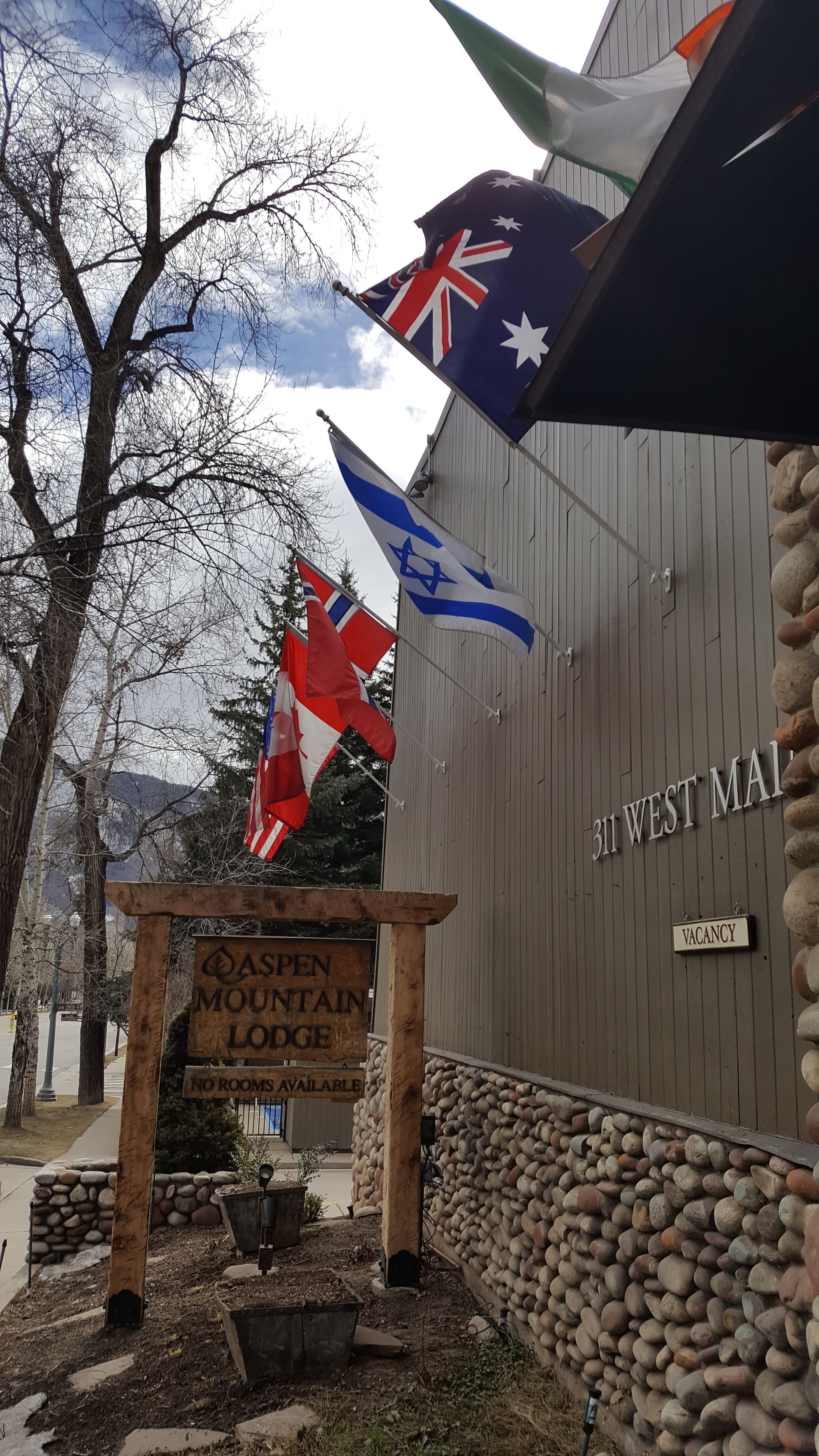 A wooden sign with "Aspen Mountain Lodge" and "No rooms available" text, flags, and a stone wall.