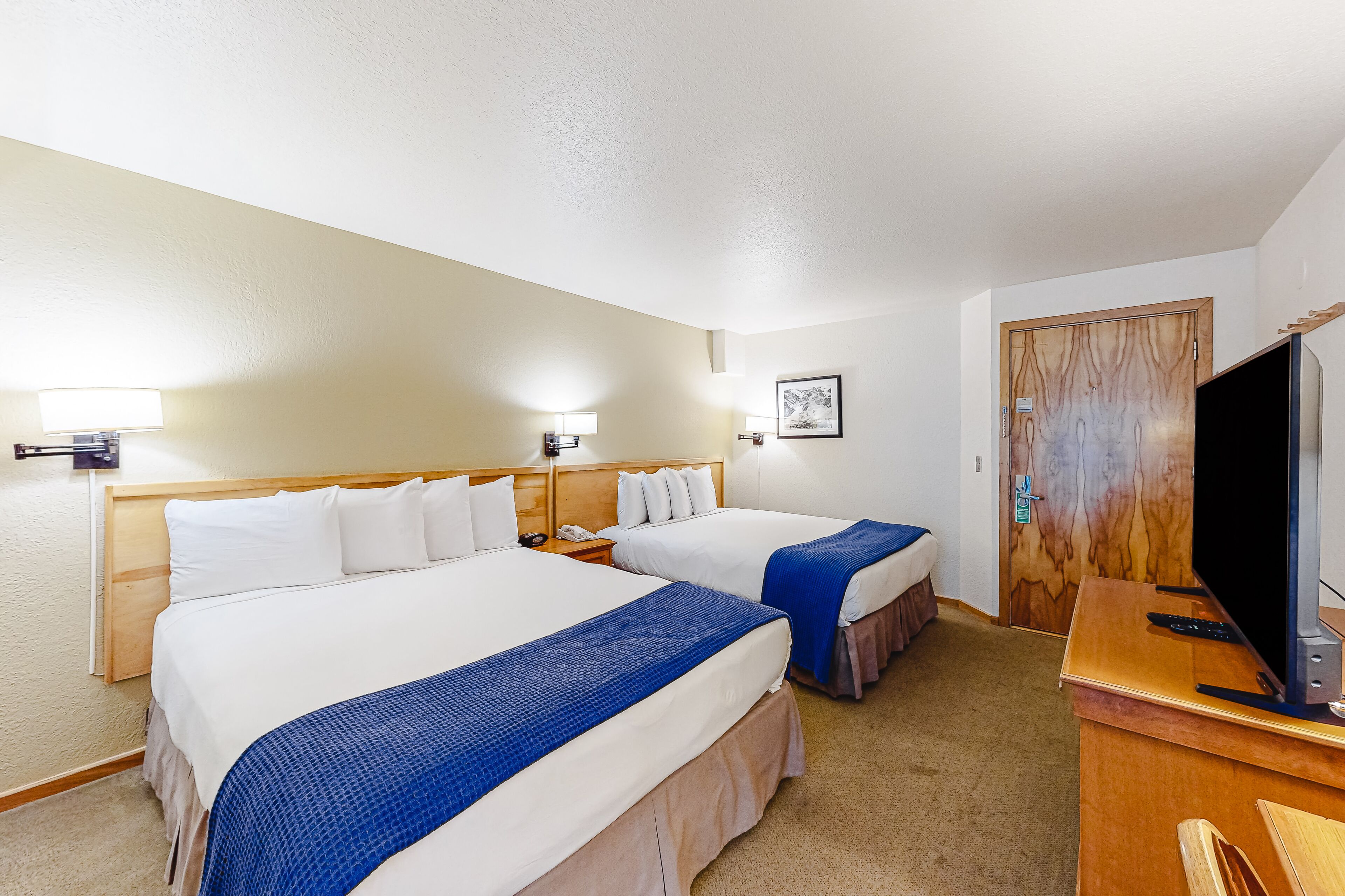 A hotel room with two beds, a desk with a television, and a door with a wooden panel.