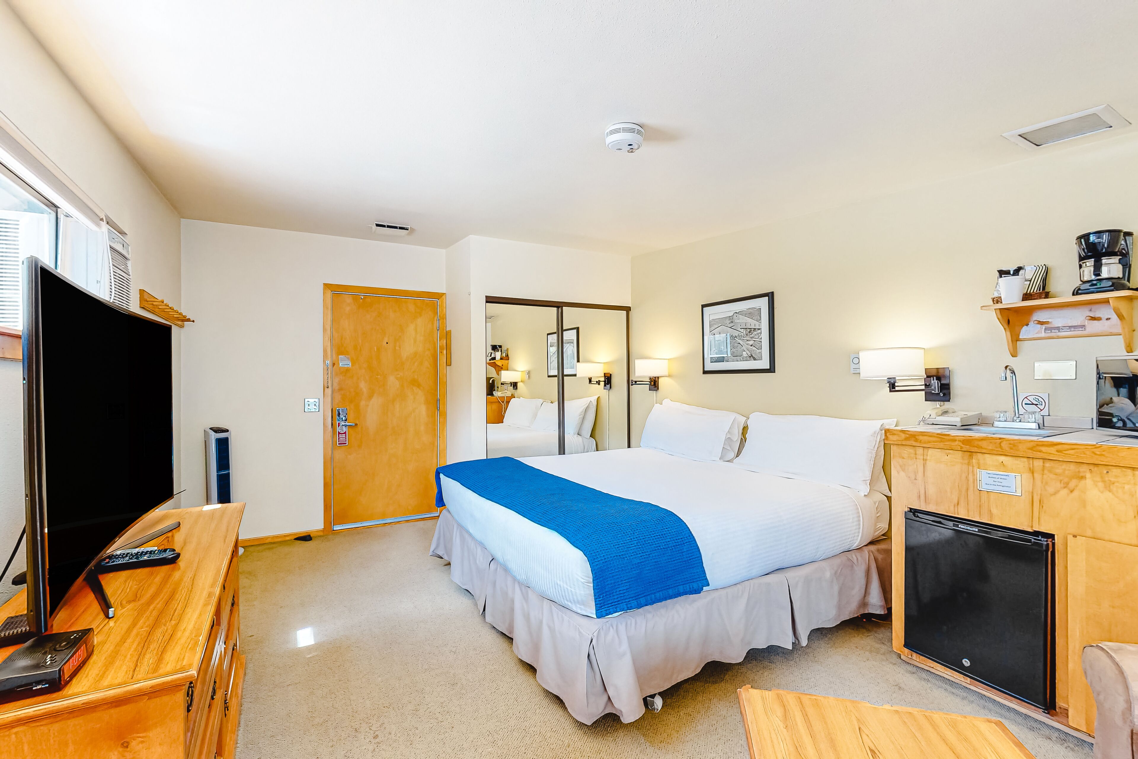 A hotel room with a bed, a desk with a TV, a refrigerator, and a wooden cabinet.