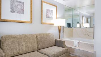 A hotel room with a sofa, a lamp, a bathtub, and two framed artworks on the wall.