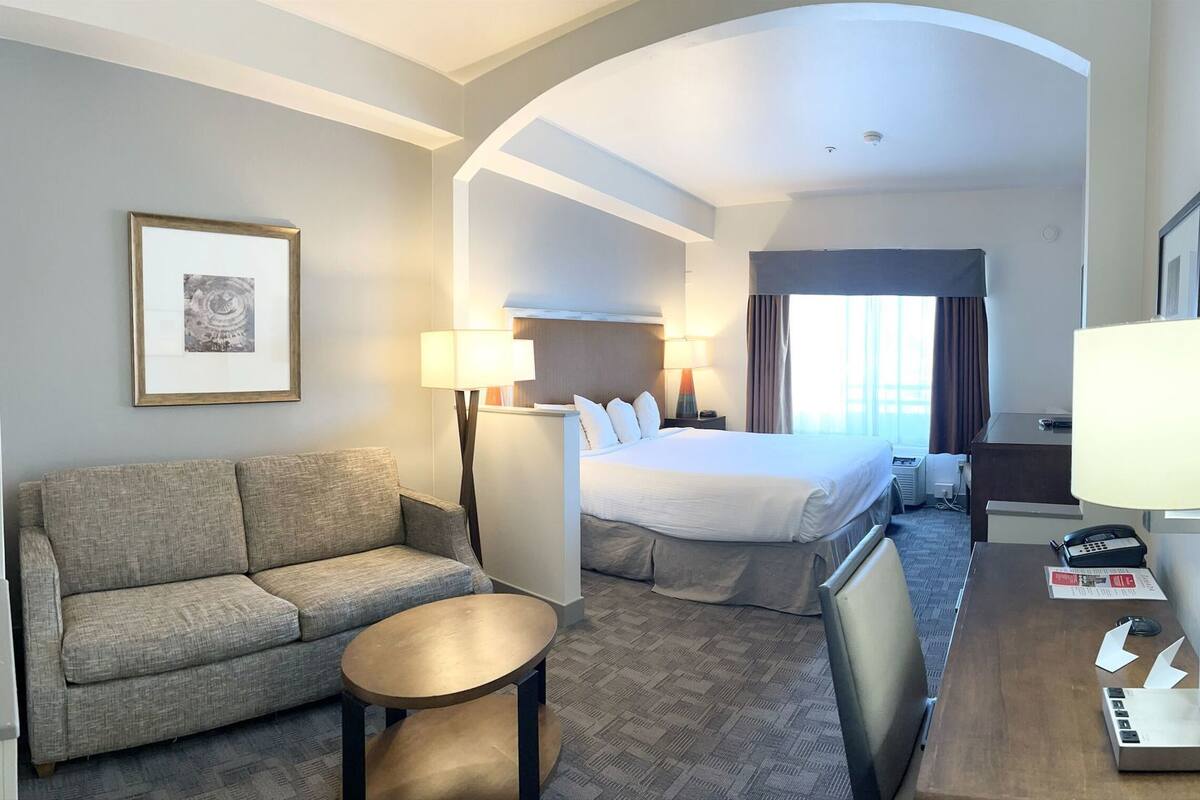 Ein Hotelzimmer mit Sofa, rundem Tisch, einem Bett mit Kissen, Schreibtisch mit Lampe und einem gerahmten Bild an der Wand.