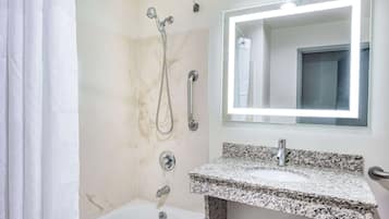 Un baño con una bañera blanca, un lavamanos de encimera de granito, un cabezal de ducha de mano y un espejo con luz.