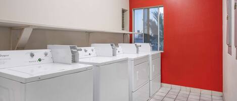 A laundry room with four white Whirlpool washers and dryers, a red wall, and a window with a view of palm trees.