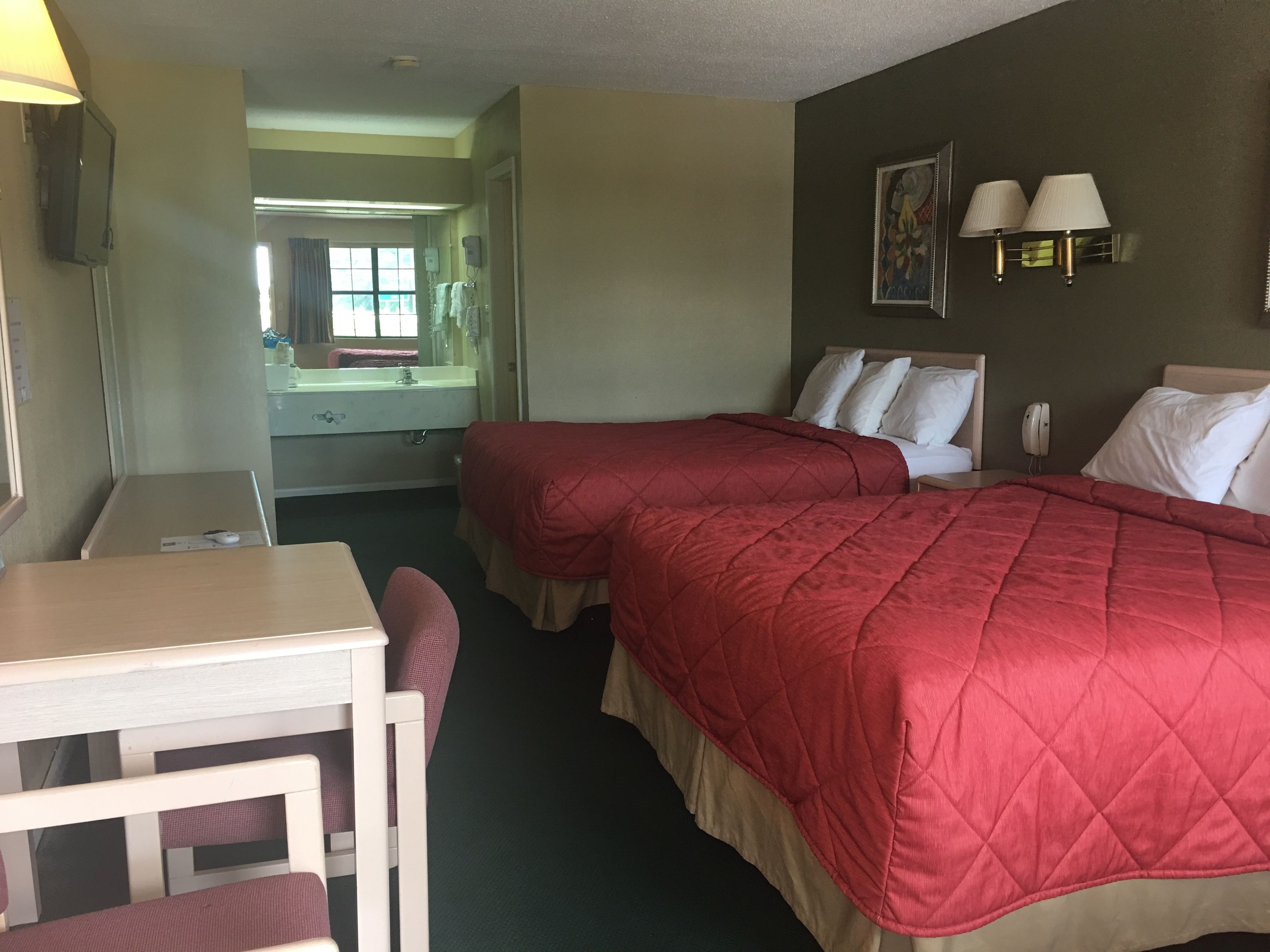 A hotel room with two beds, a desk, a chair, a mirror, a sink, and a wall-mounted lamp.