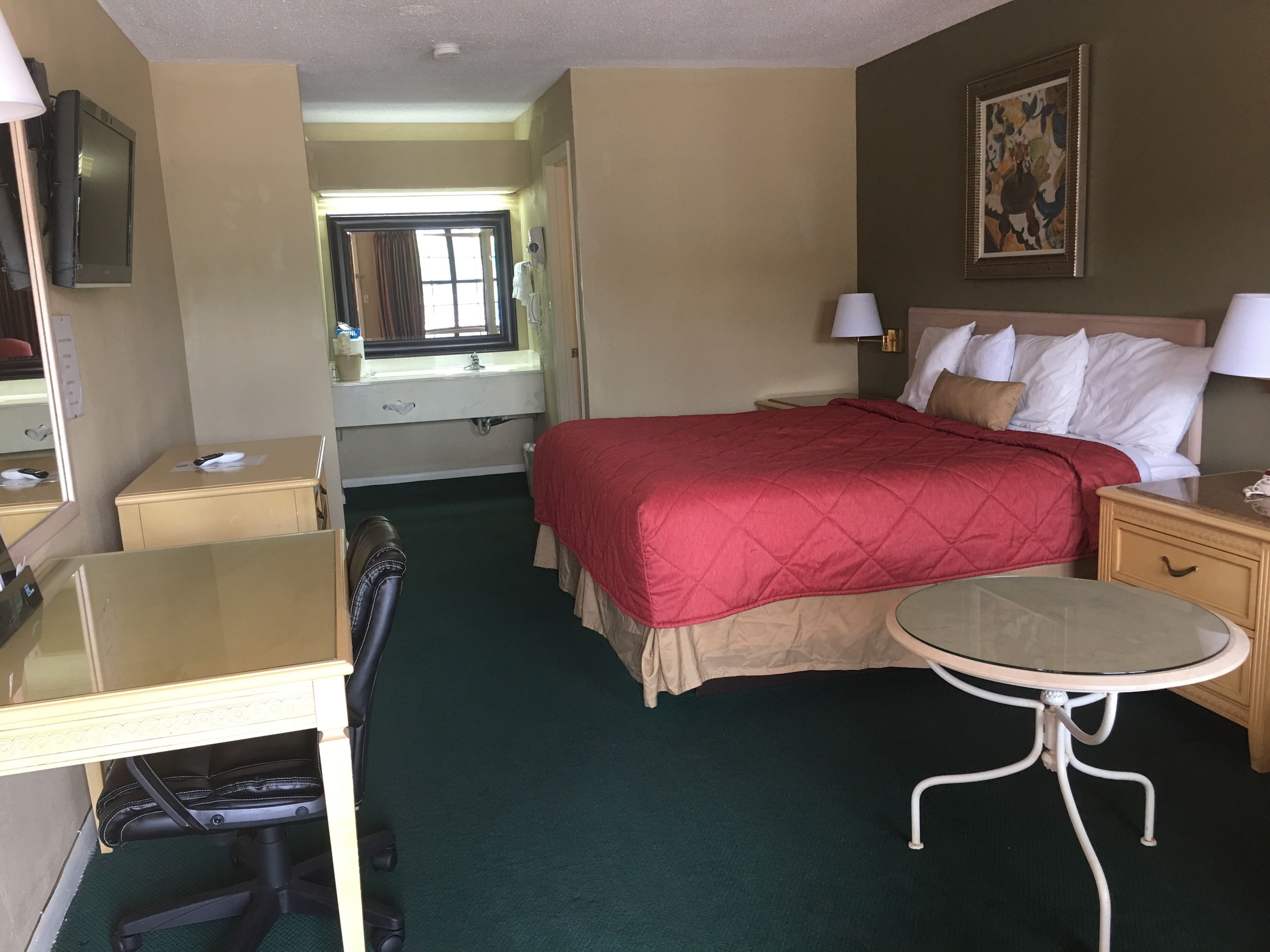 Una habitación de hotel con cama, escritorio, silla, mesita, televisor y un cuadro en la pared.