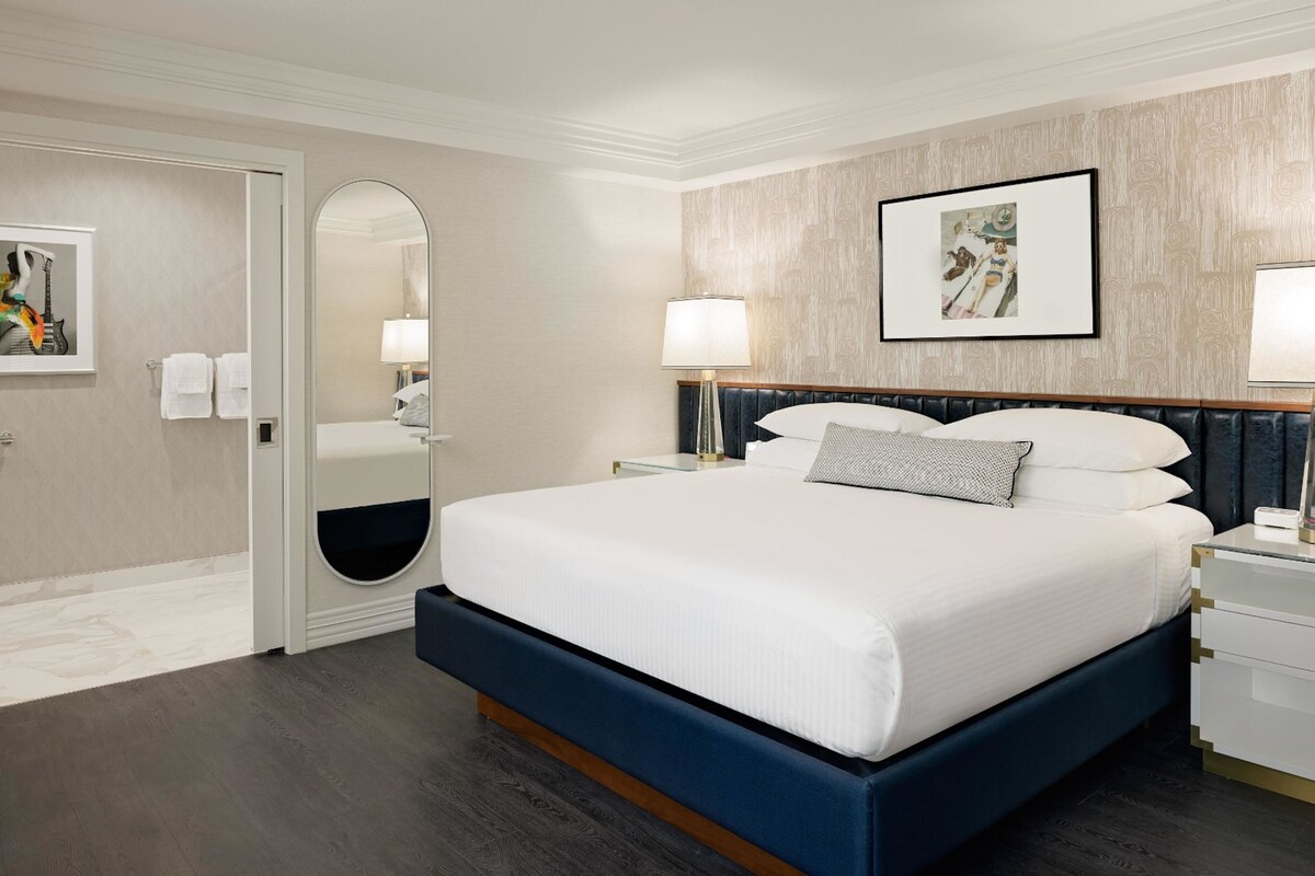 Une chambre d’hôtel moderne avec un grand lit, des tables de chevet, un miroir et des œuvres d’art accrochées aux murs.