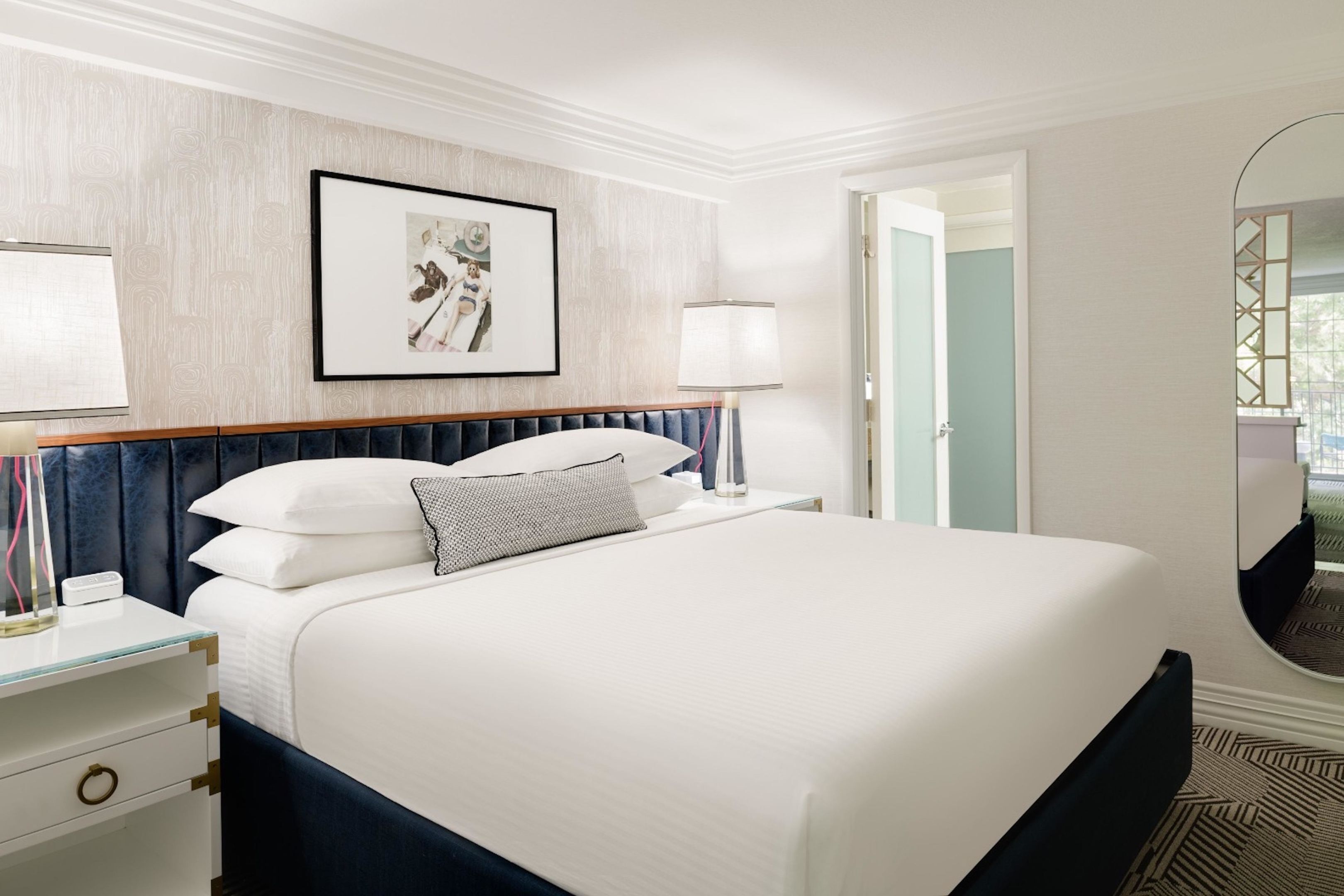 A hotel room with a bed, bedside table, lamp, framed picture, and a mirror.