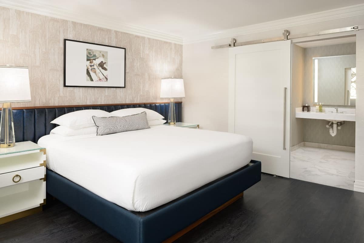 A modern hotel room with a large bed, bedside lamps, a framed artwork, a walk-in closet, and a bathroom with a mirror and sink.