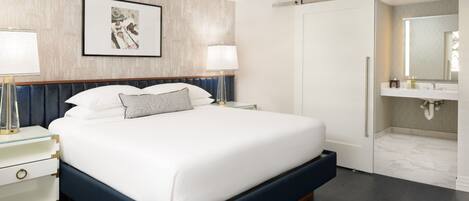 Una habitación de hotel moderna con una cama grande, lámparas de noche, una obra de arte enmarcada, un vestidor y un baño con espejo y lavamanos.