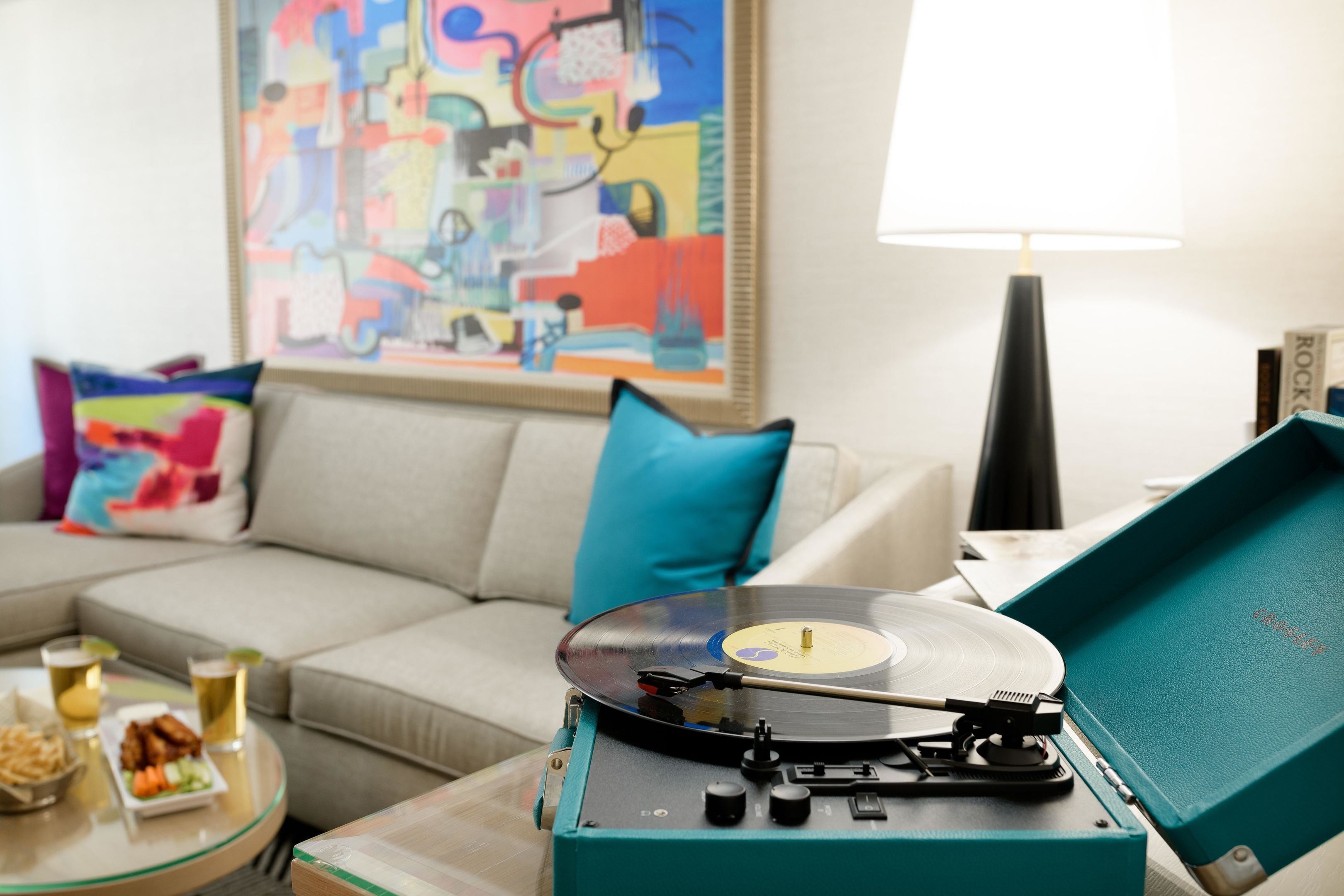A living room with a teal record player, a beige sofa, a colorful abstract painting, and a lamp.