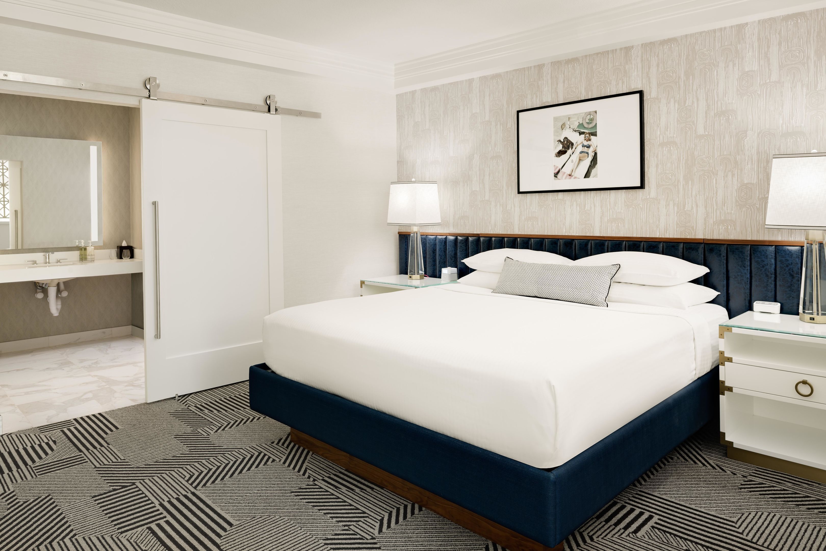 Une chambre d’hôtel avec un lit, des tables de chevet, un miroir, un tableau encadré au mur et un tapis à motifs.