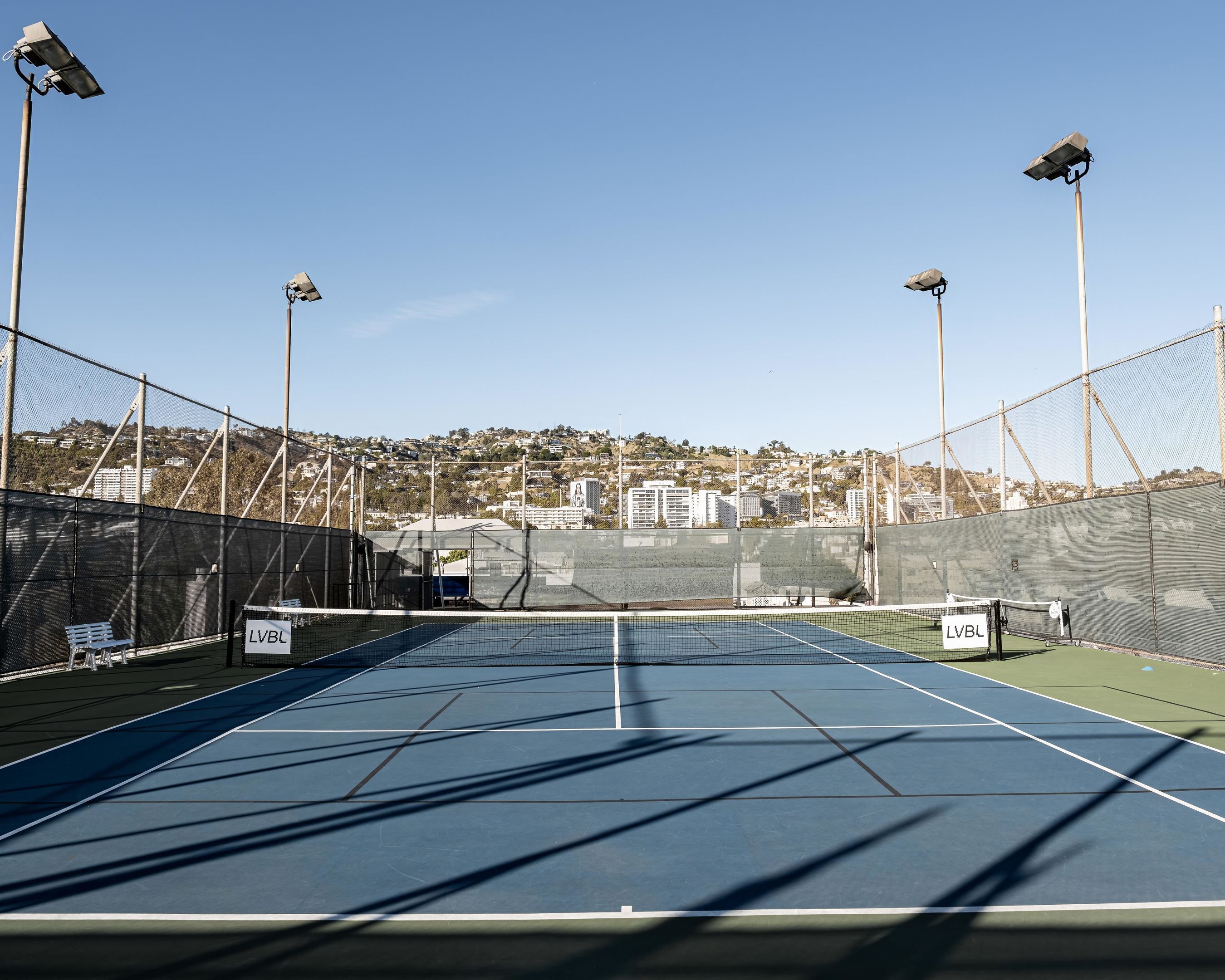 An outdoor tennis court with net, surrounded by fencing, and equipped with floodlights.