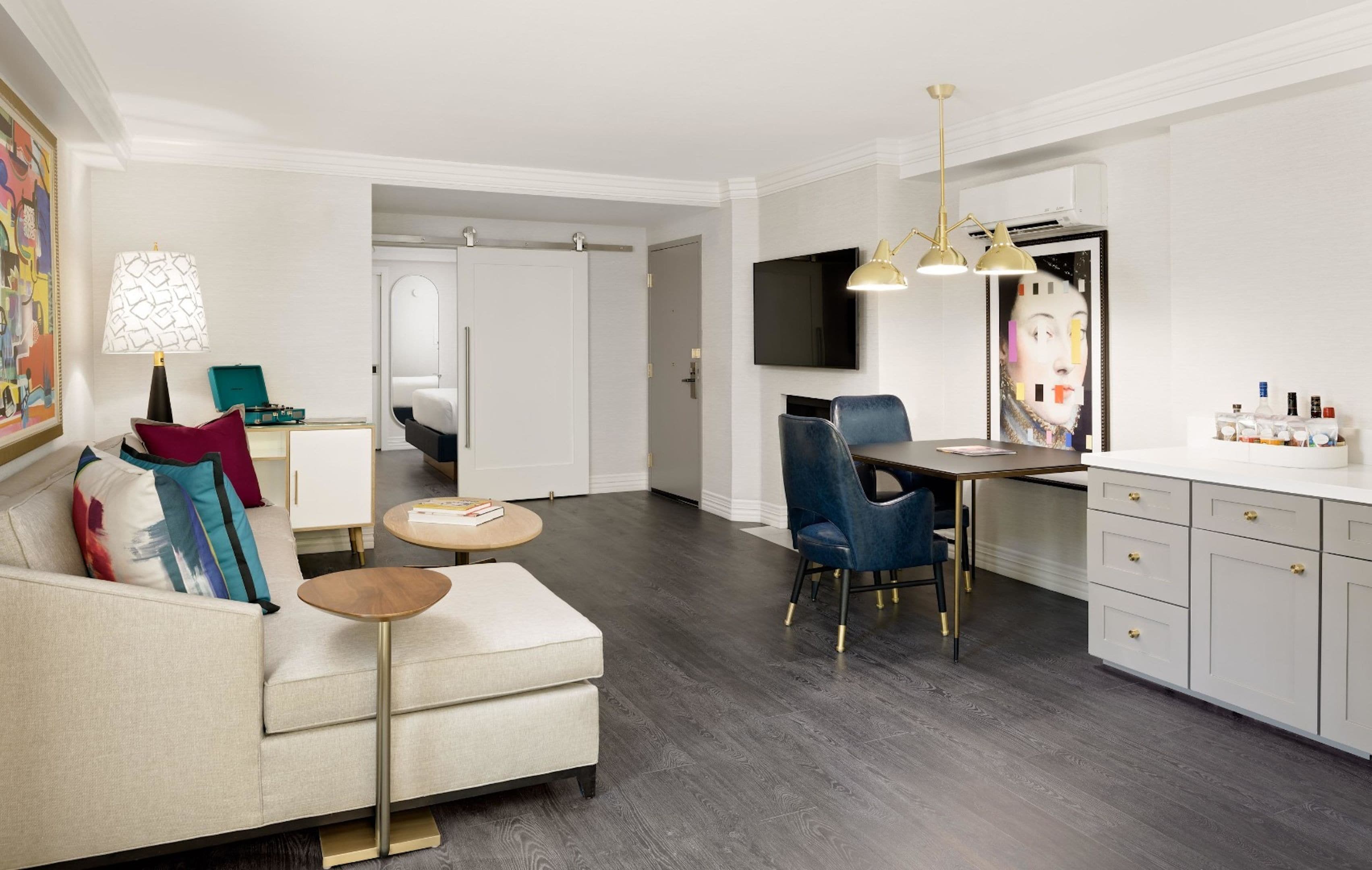 A modern hotel room with a sofa, a small round table, a desk with a chair, a television, and a cabinet with drawers.