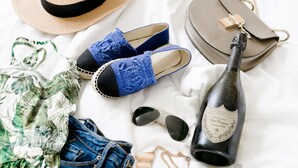 A bed with a straw hat, blue lace slippers, a grey handbag, a bottle of champagne, sunglasses, a floral print bikini, and denim shorts.