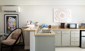 A modern kitchen with a granite countertop, stainless steel sink, and a microwave. There is a chair with a striped cushion, a framed artwork on the wall, and a decorative wall piece.