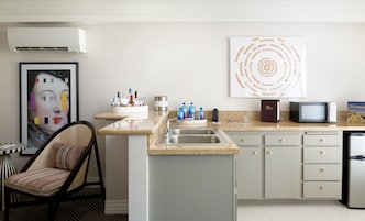A modern kitchen with a granite countertop, stainless steel sink, and a microwave. There is a chair with a striped cushion, a framed artwork on the wall, and a decorative wall piece.