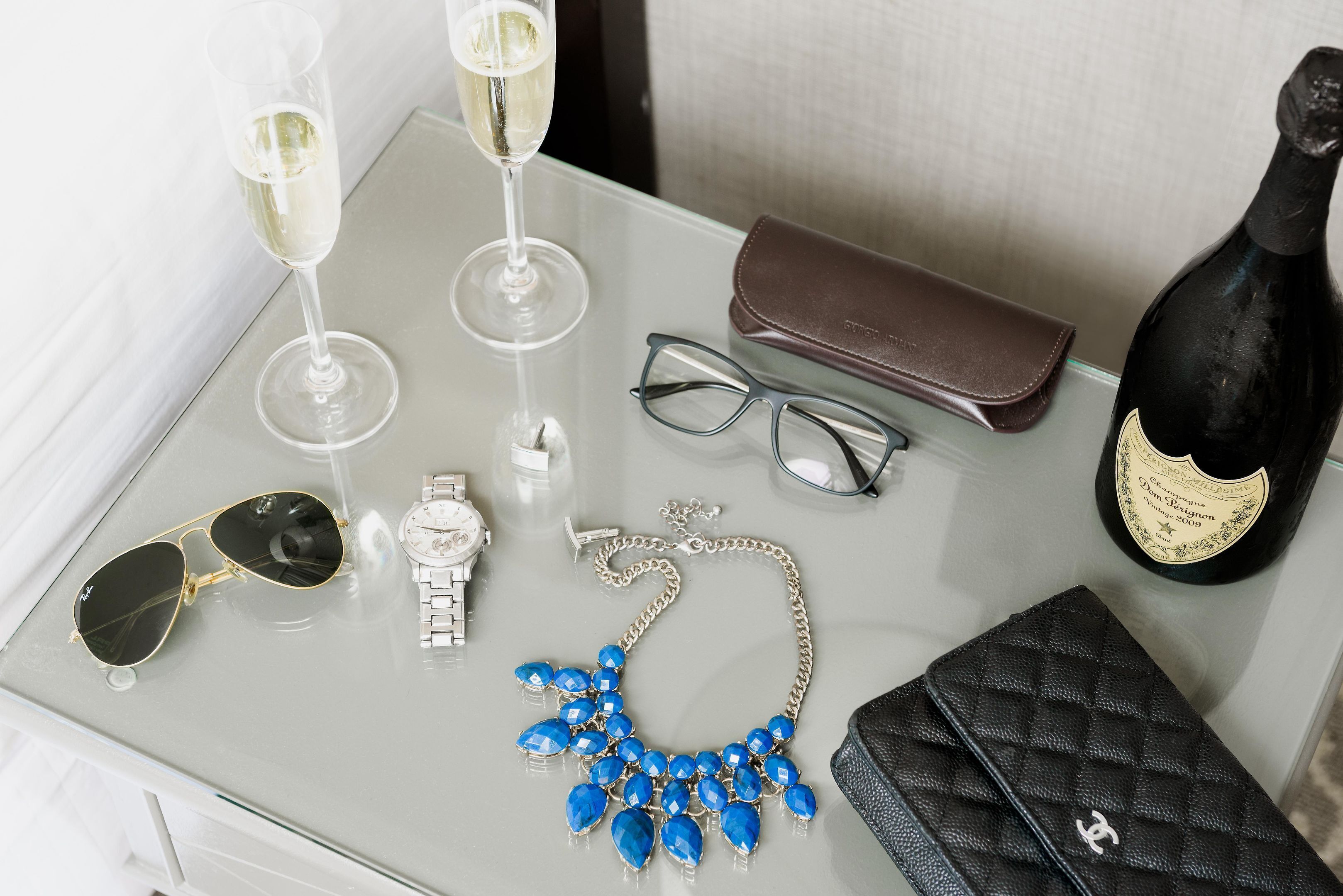 A glass table with a watch, sunglasses, a blue necklace, a black handbag, a pair of glasses, and two champagne flutes.