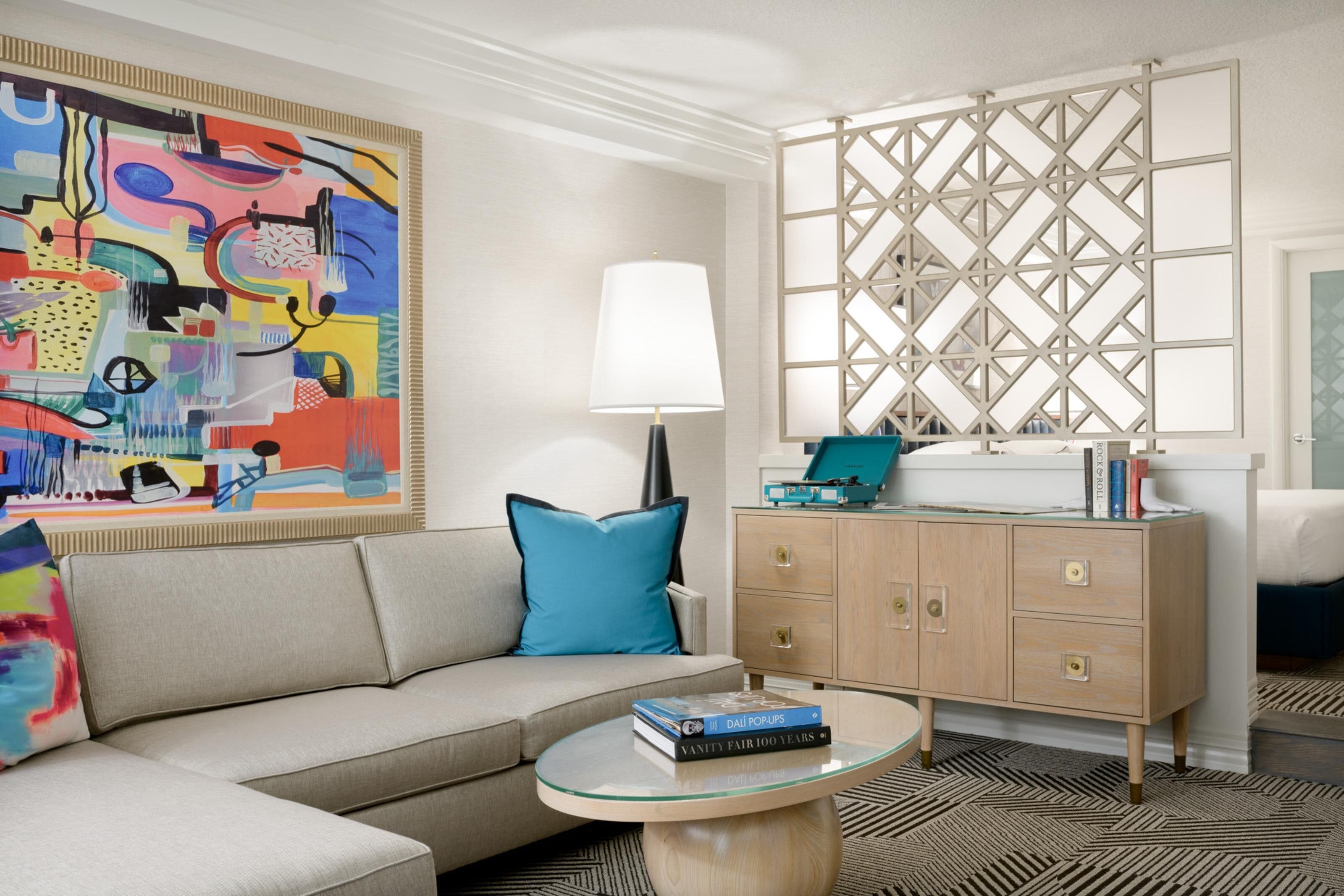 A modern living room with a sofa, a glass coffee table, a decorative lamp, a cabinet with books, and a colorful abstract painting.