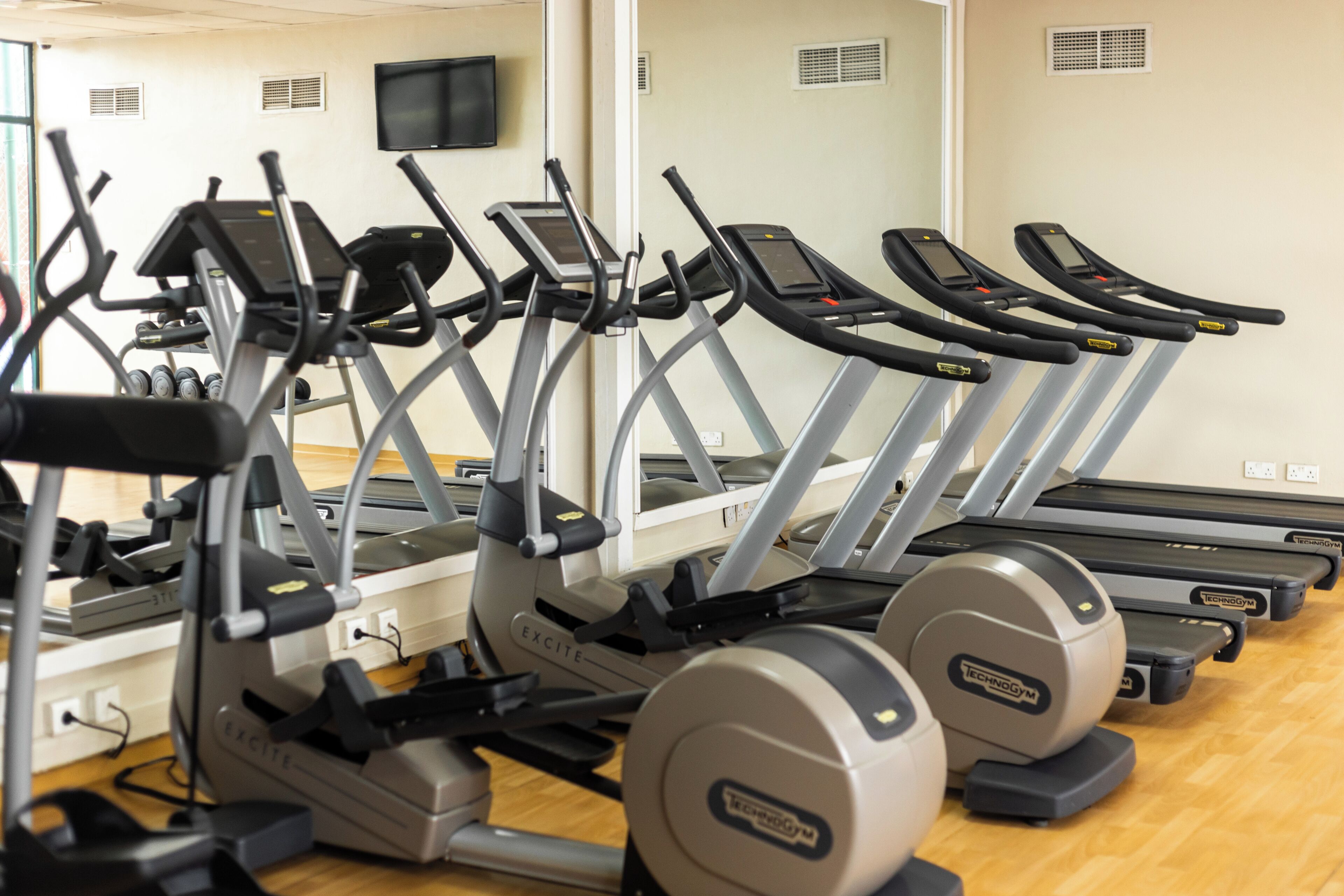 A row of treadmills and elliptical machines in a gym with a wooden floor and white walls.