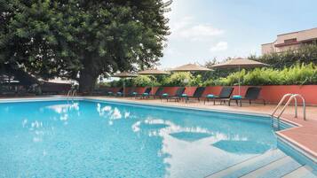 Piscina con agua azul cristalina, rodeada de sillas de descanso y sombrillas, con un árbol grande que da sombra.