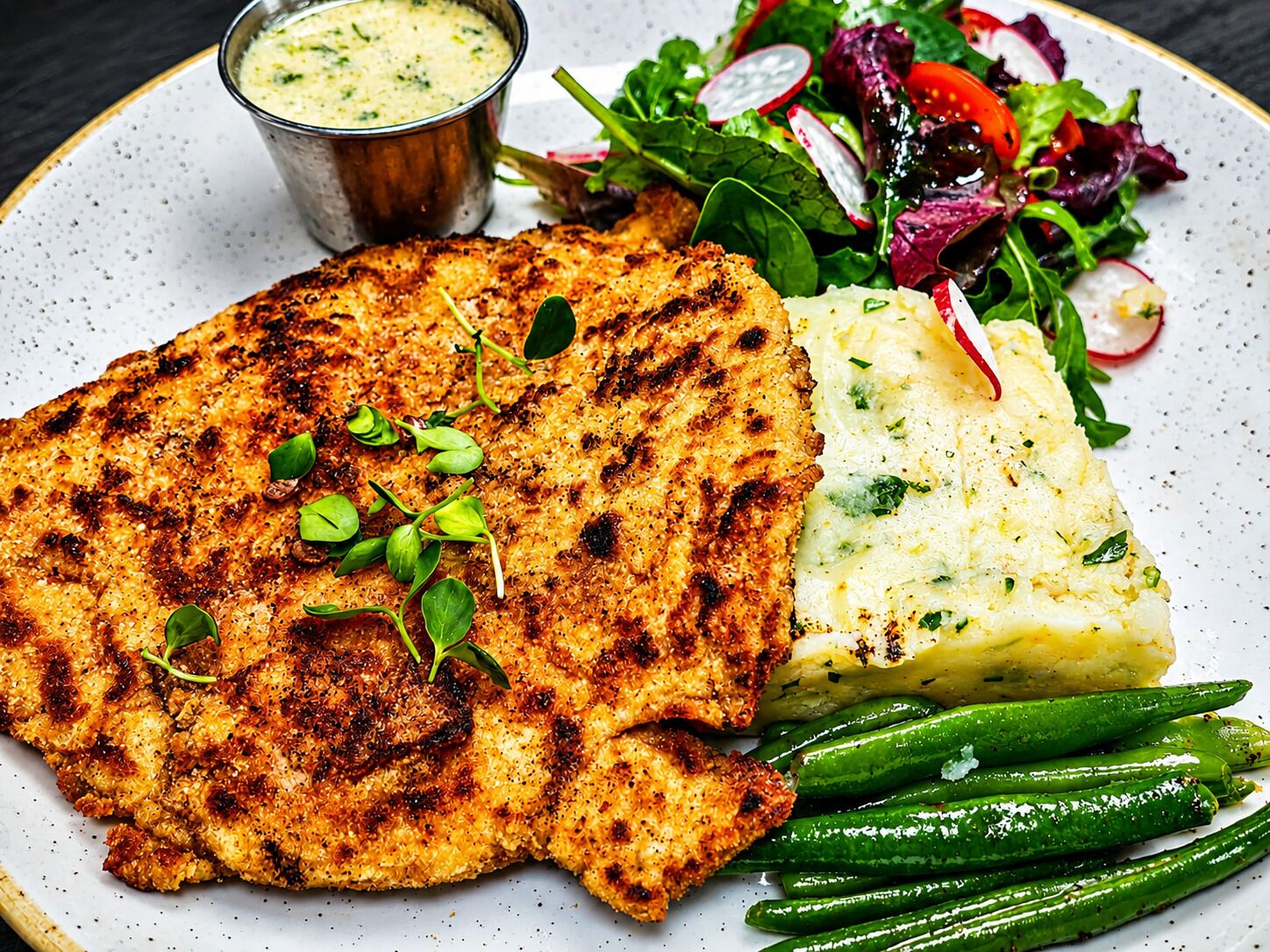Une assiette composée de viande grillée, de purée de pommes de terre, de haricots verts et d’une salade de radis.
