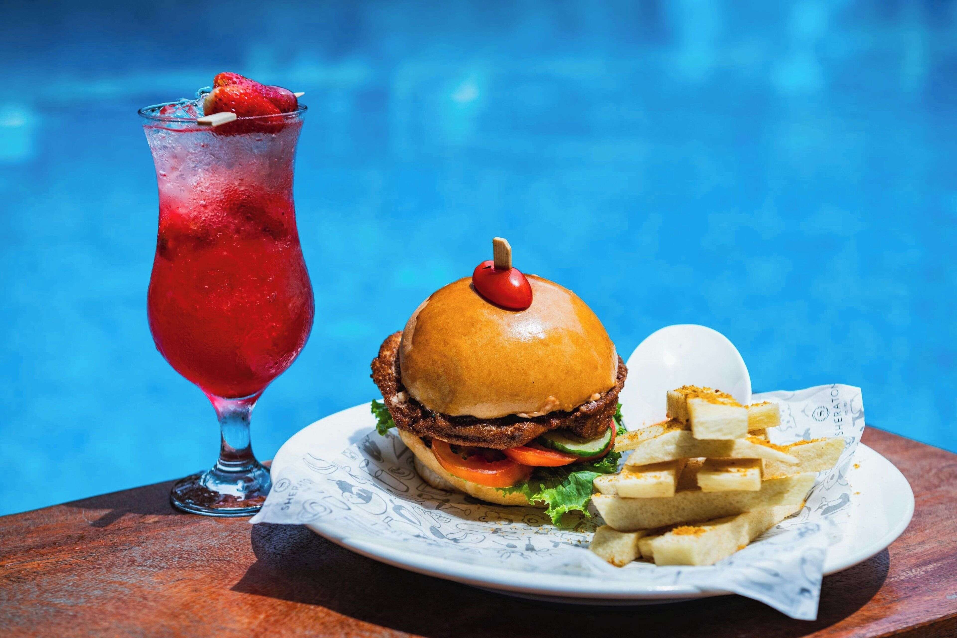 A burger with a cherry on top, a drink with a strawberry, and fries on a plate by a pool.