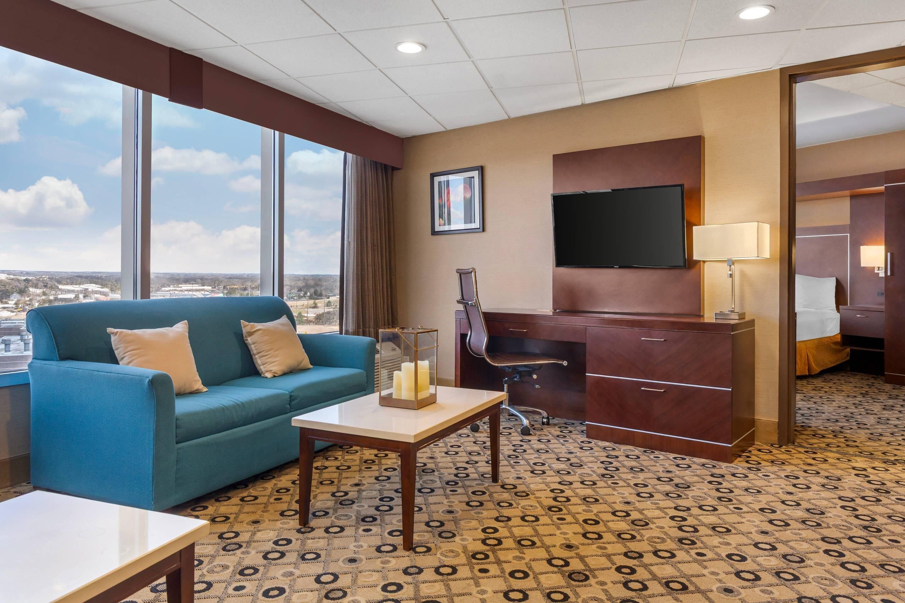 A hotel room with a large window, a blue sofa, a desk with a chair, and a television.