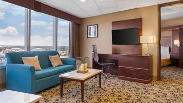A hotel room with a large window, a blue sofa, a desk with a chair, and a television.