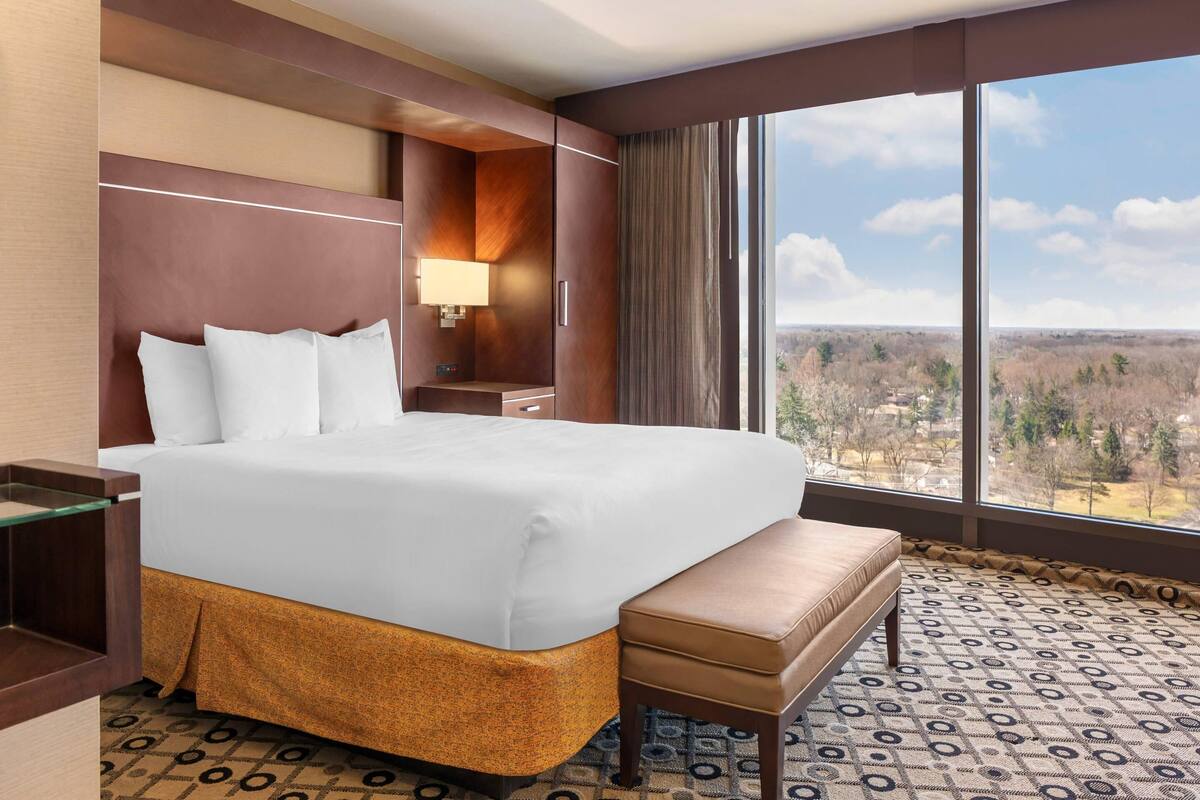 Une chambre d’hôtel avec un grand lit, un banc, une table de chevet, une lampe et une grande fenêtre donnant sur le paysage.