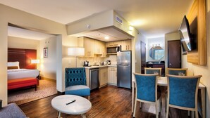 A compact hotel room with a kitchenette, a dining area with blue upholstered chairs, a small round table, a bed with a red accent, and a TV mounted on the wall.