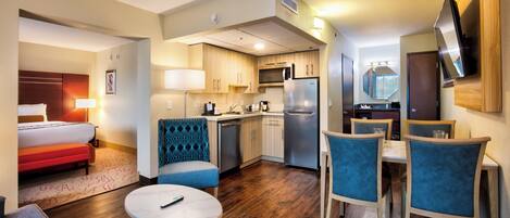 A compact hotel room with a kitchenette, a dining area with blue upholstered chairs, a small round table, a bed with a red accent, and a TV mounted on the wall.