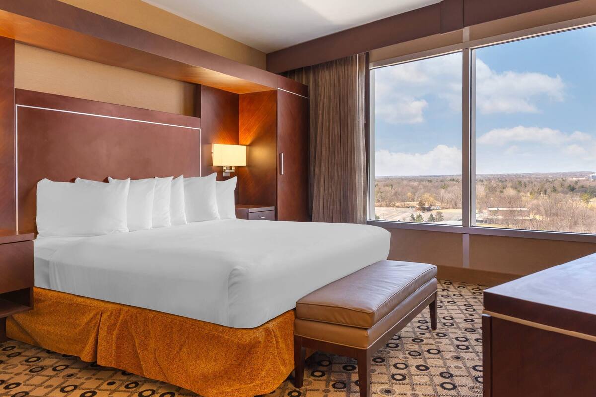 Une chambre d’hôtel dotée d’un grand lit, d’un bureau, d’une banquette et d’une grande fenêtre donnant sur le paysage.