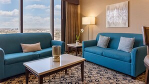 A hotel room with two blue sofas, a small table, a lamp, a painting, and a view of the city through large windows.
