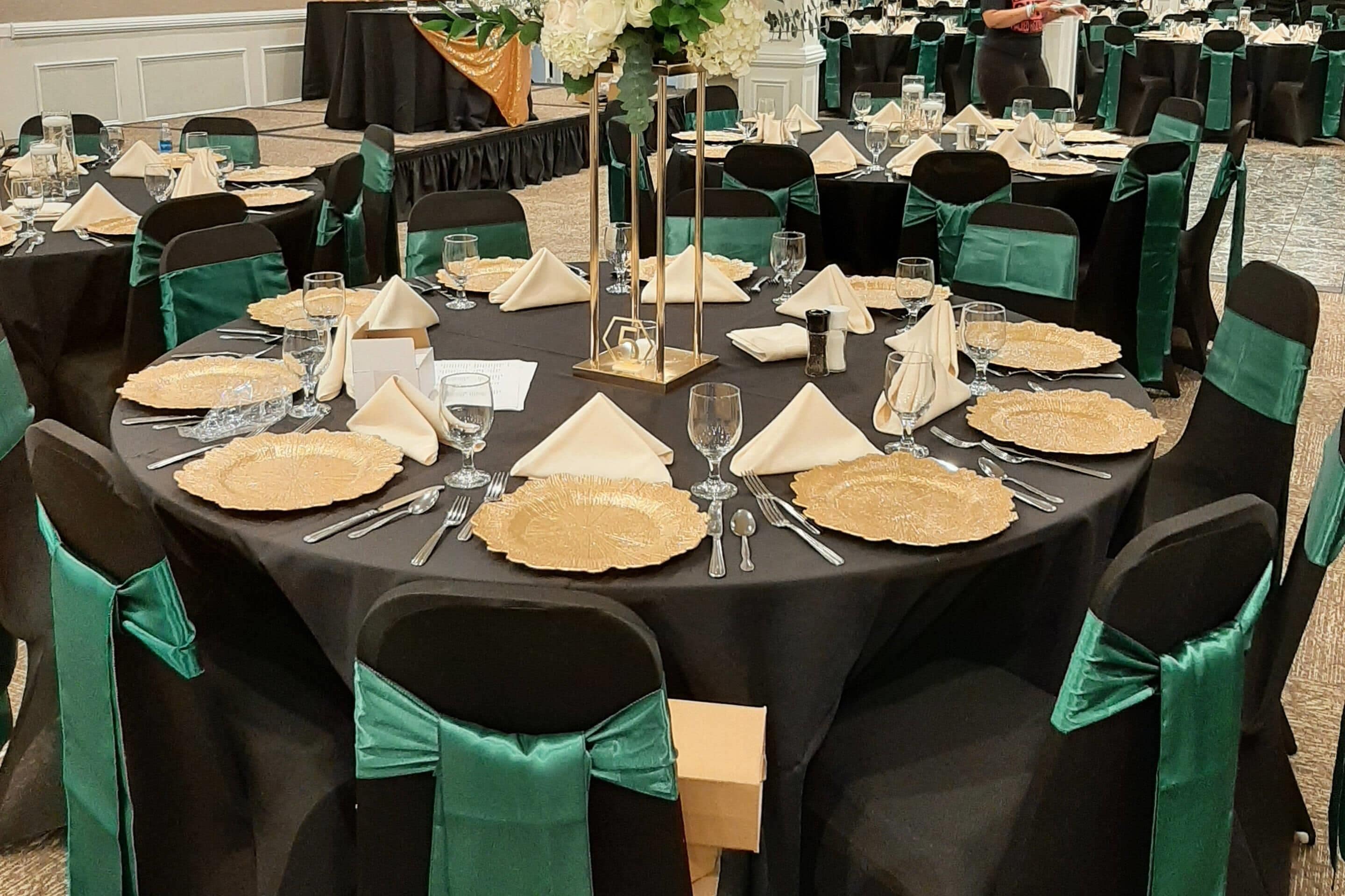 Un salón de banquetes con mesas redondas dispuestas para un evento formal, cada una con un plato con borde dorado, cubiertos y una servilleta doblada.