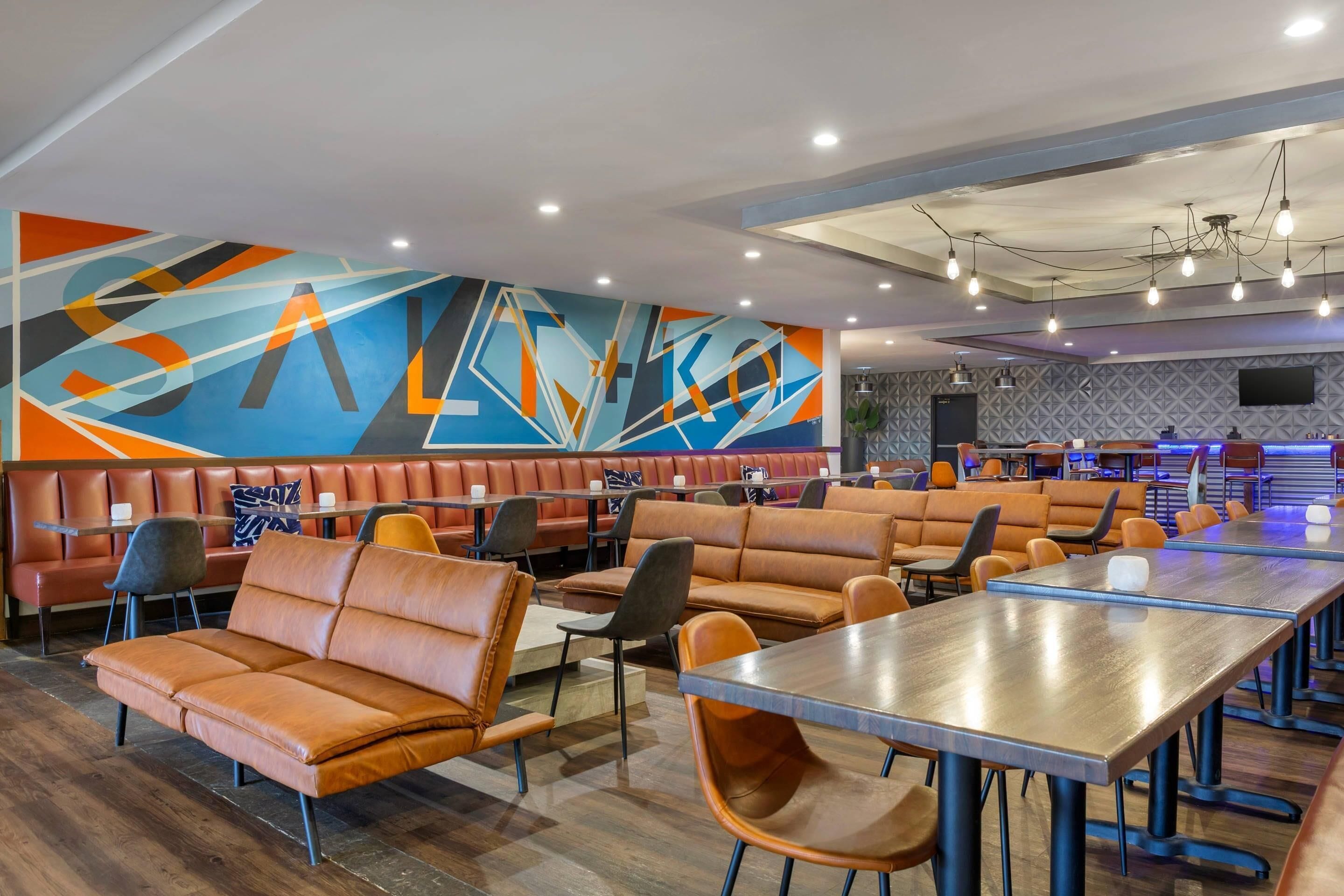 A modern restaurant with leather booths and wooden tables, a colorful abstract mural on the wall, and a flat-screen TV mounted on the wall.