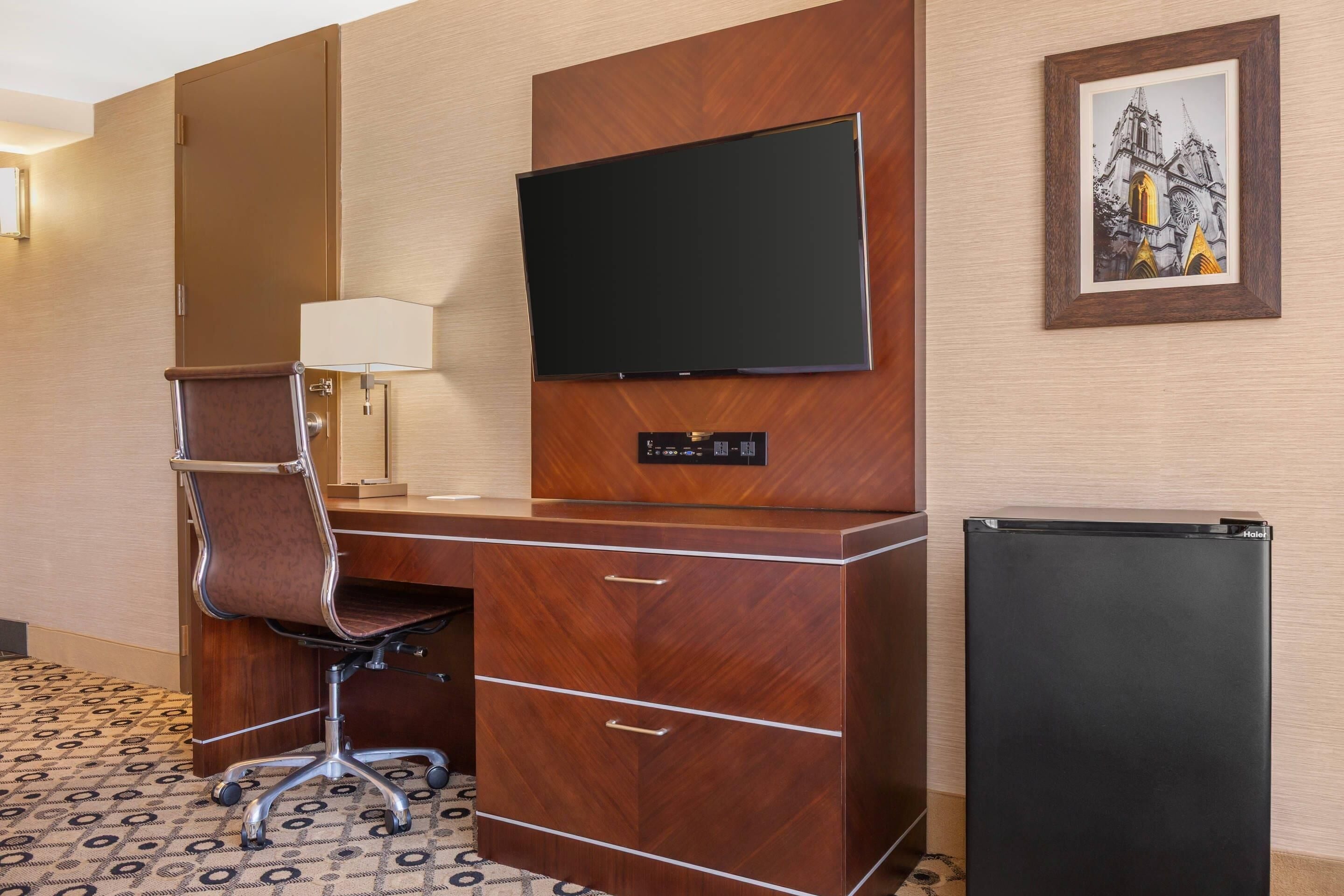 A hotel room with a wooden desk, a chair, a flat-screen TV mounted on the wall, a small refrigerator, and a framed picture above the TV.