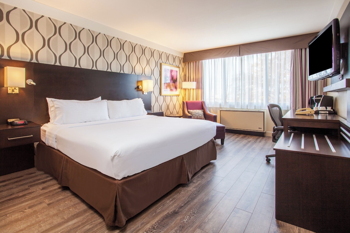 A hotel room with a large bed, a desk with a chair, a flat-screen TV, and a window with curtains.
