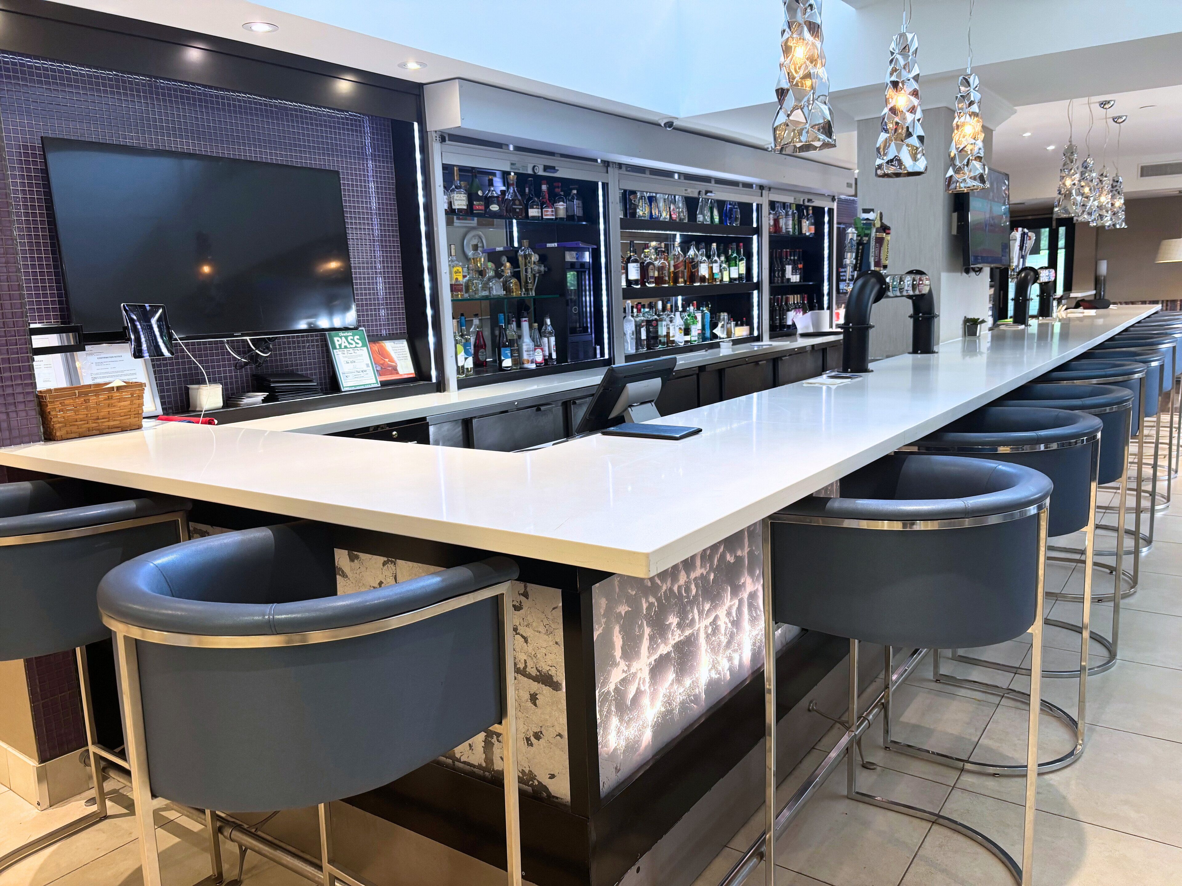 Zona de bar moderna con una barra alargada, varios taburetes y una amplia variedad de botellas de licor.