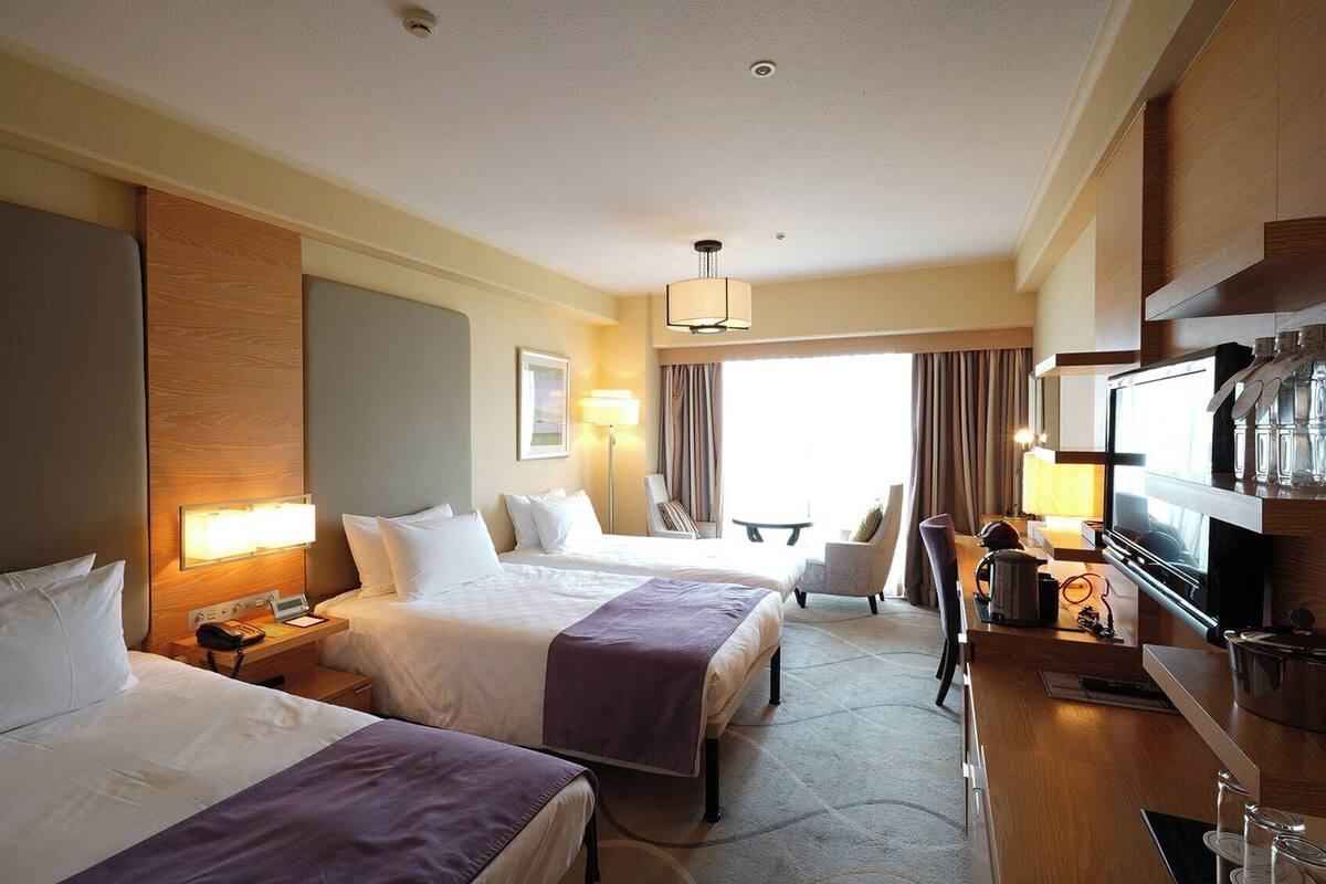 A hotel room with two beds, a desk with a television, a chair, and a large window with curtains.