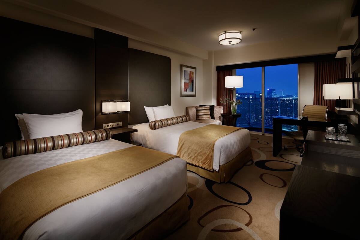 A hotel room with two beds, a desk, and a view of the city at night.