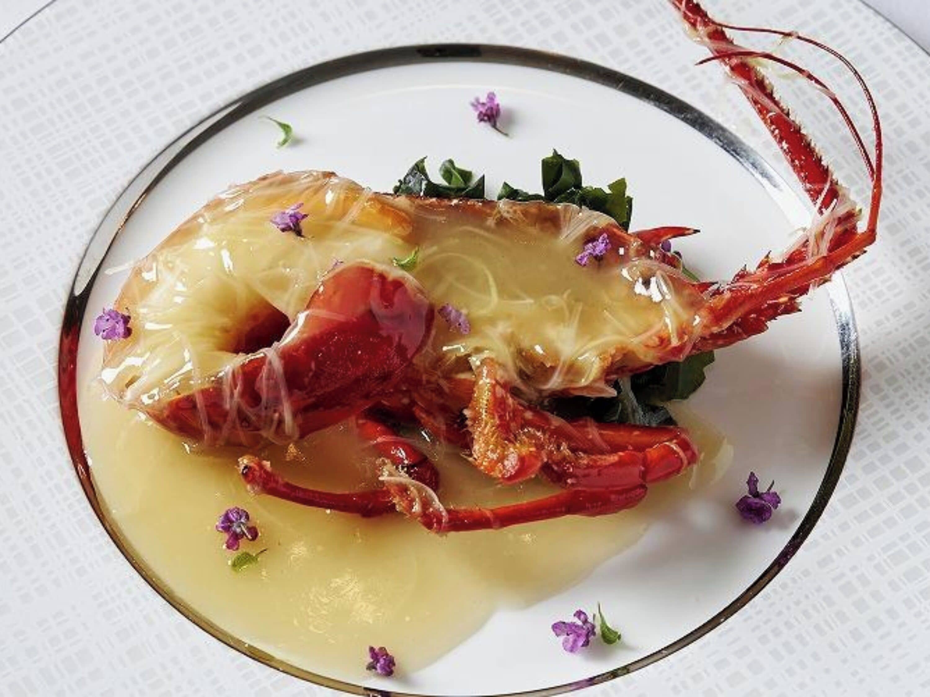 A lobster dish on a plate with a clear liquid and garnished with purple flowers.