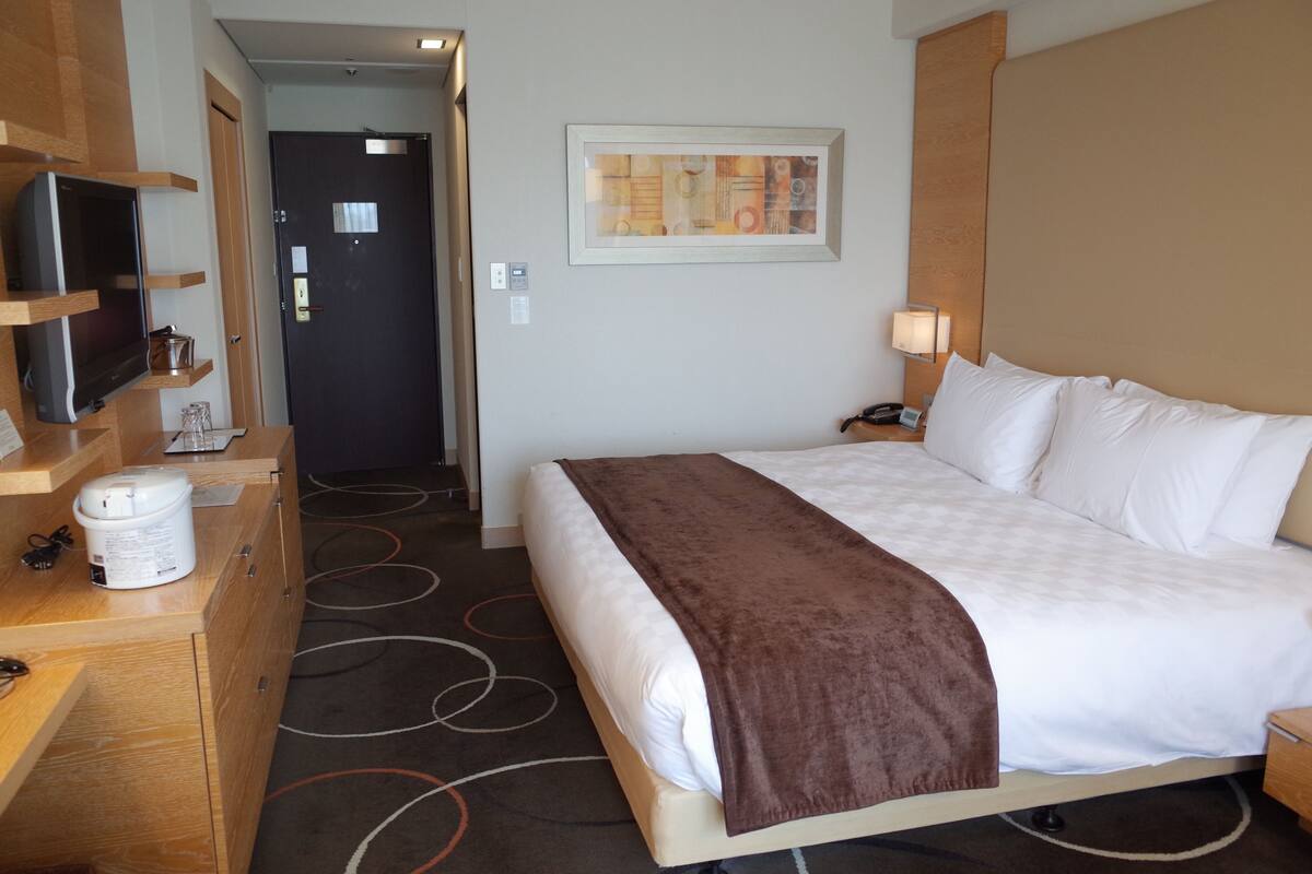 A hotel room with a bed, a television on a shelf, a desk with a coffee maker, and a door leading to another room.