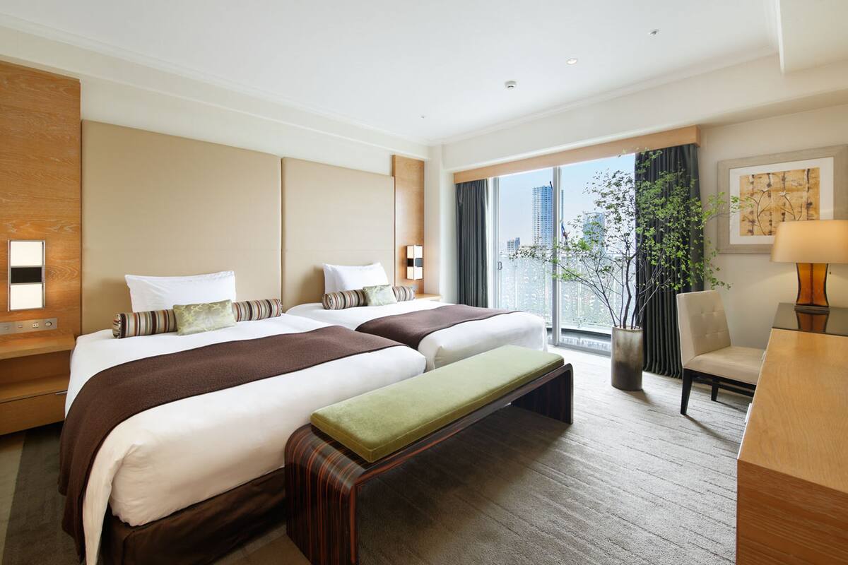 A hotel room with two beds, a bench, a desk, a chair, a potted plant, and a large window with a city view.
