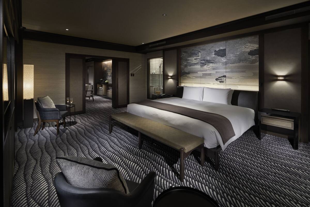 A modern hotel room with a large bed, a bench, a desk, and a chair. There is a wall-mounted TV and a lamp.