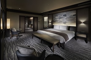 A modern hotel room with a large bed, a bench, a desk, and a chair. There is a wall-mounted TV and a lamp.